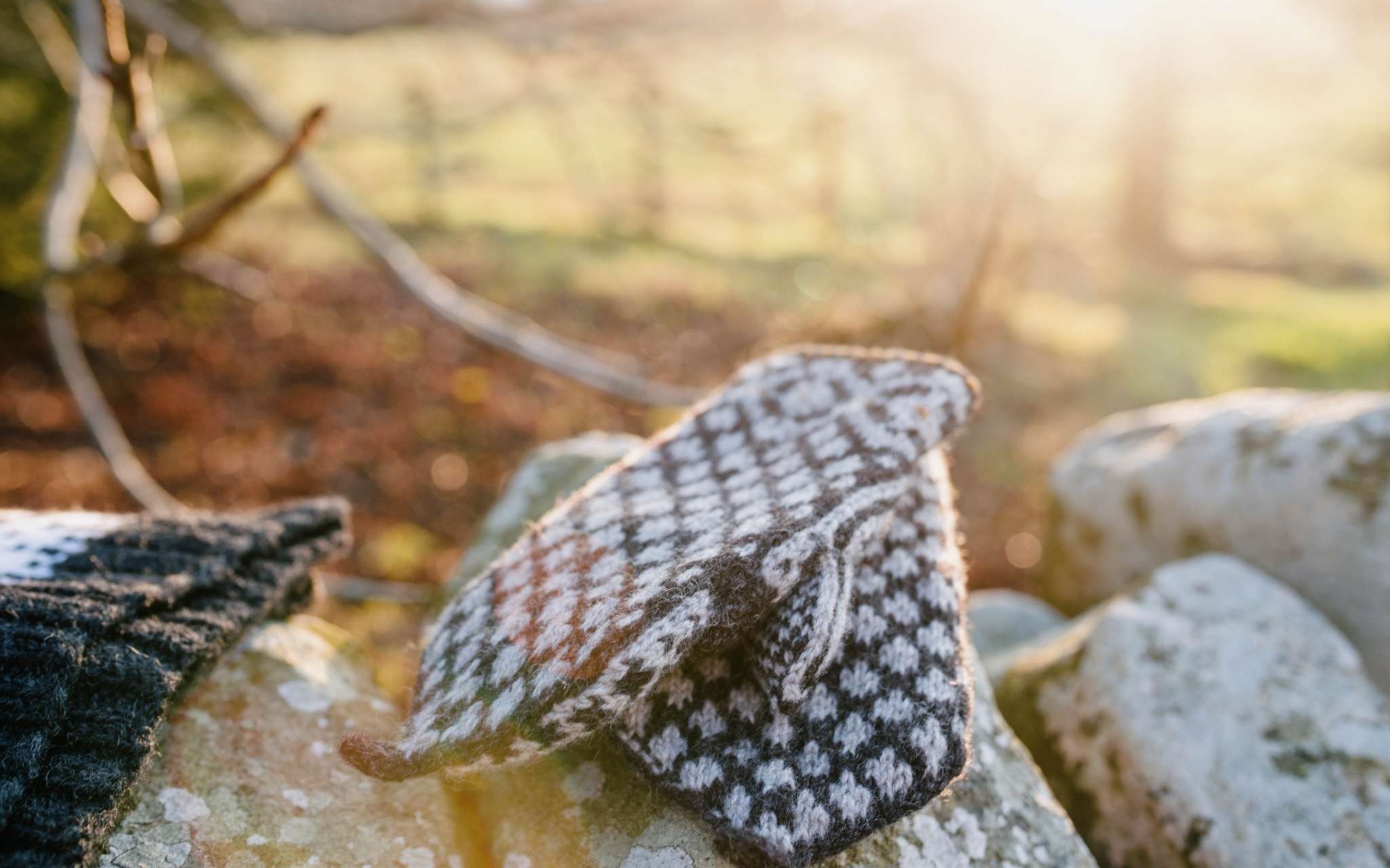 A pair of dark and cream colourwork mittens lay on top of each other, on a stone wall. The light shines in the background through a fence.