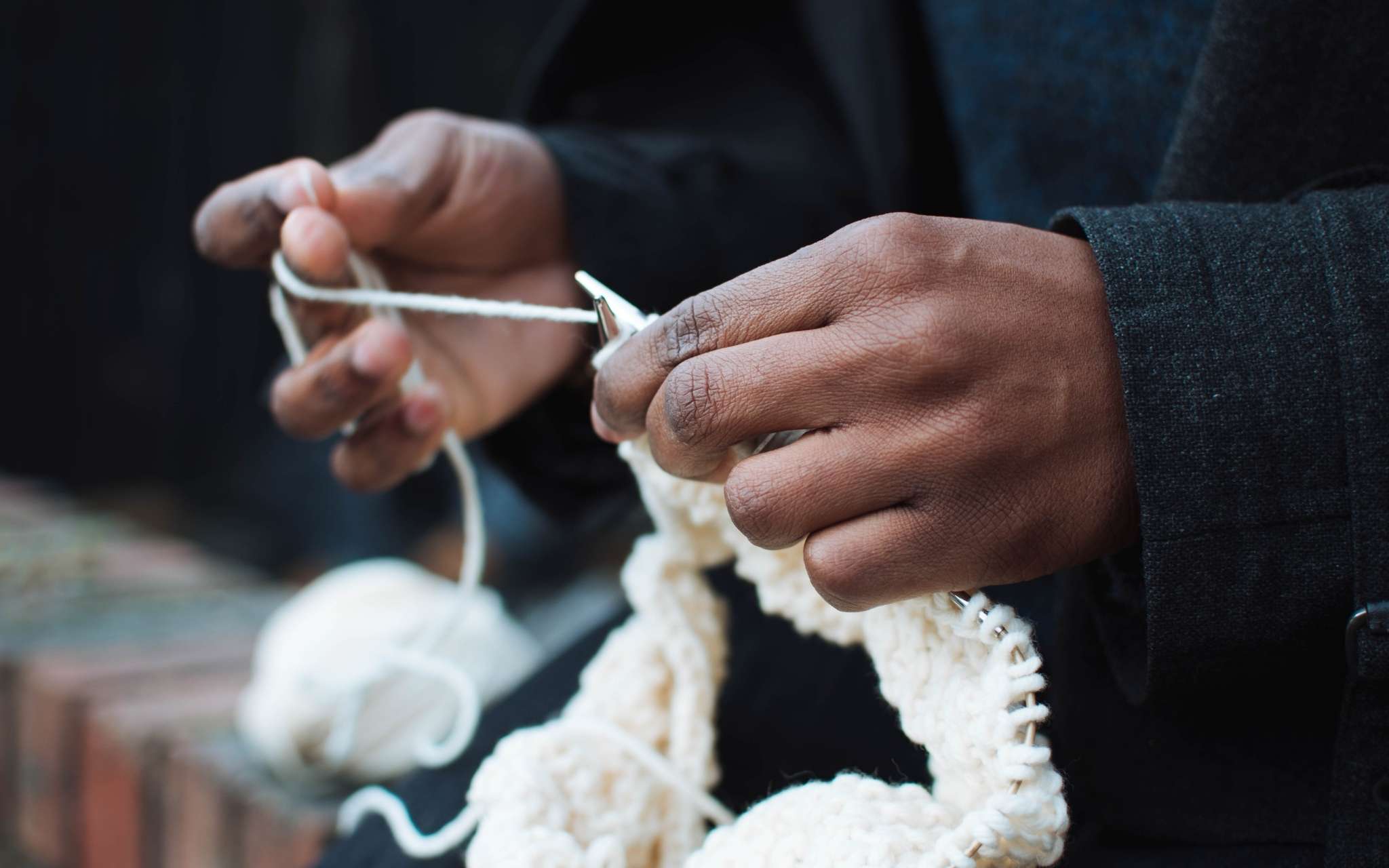 A black person knits on a white sweater with circular needs, the ball of yarn lies on a wall to the side.