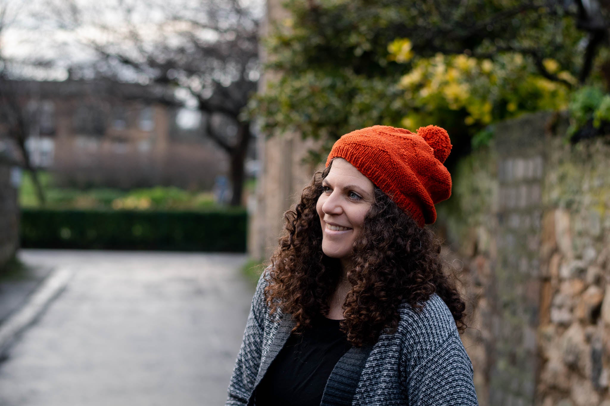 A woman with white skin and dark curly hair stands in a street beside a brick wall. She is wearing a bright orange hat with pom pom and open grey jacket and is smiling.