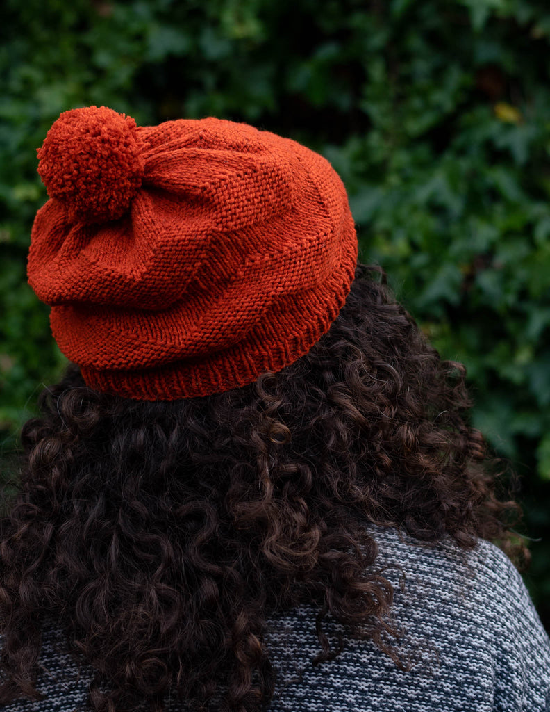 close up of the back of a beanie on a person with long curly hair, the beanie is burnt orange with a textured chevron pattern and a pom pom