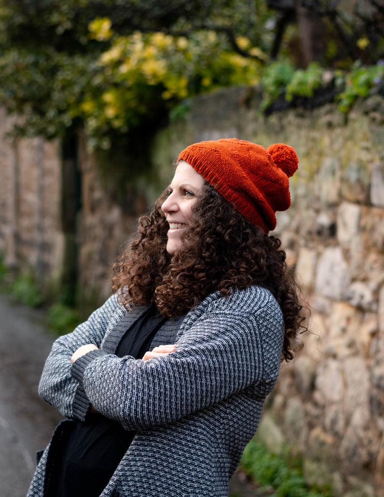 a person with long curly hair standing with their arms folded wearing a slouchy beanie , the beanie is burnt orange with a textured chevron pattern and a pom pom