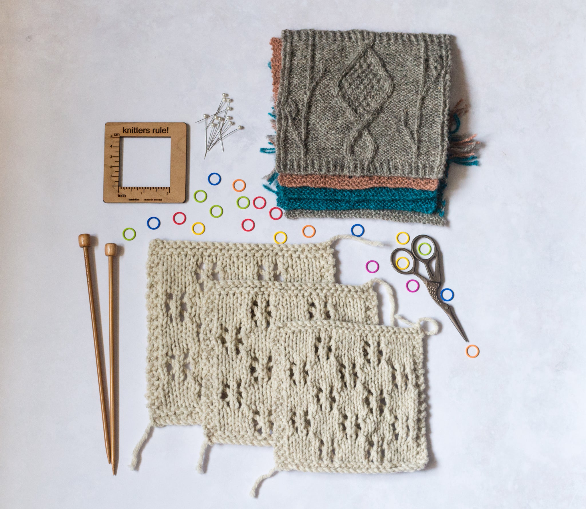Three natural coloured lace swatches like overlapping each other, with a pile of swatches above in the top right of the image. Also arranged next to the swatches in a pair straight wooden knitting needles, a square wooden measuring tool, t-pins, brightly coloured stitch markers and a small pair of scissors. 