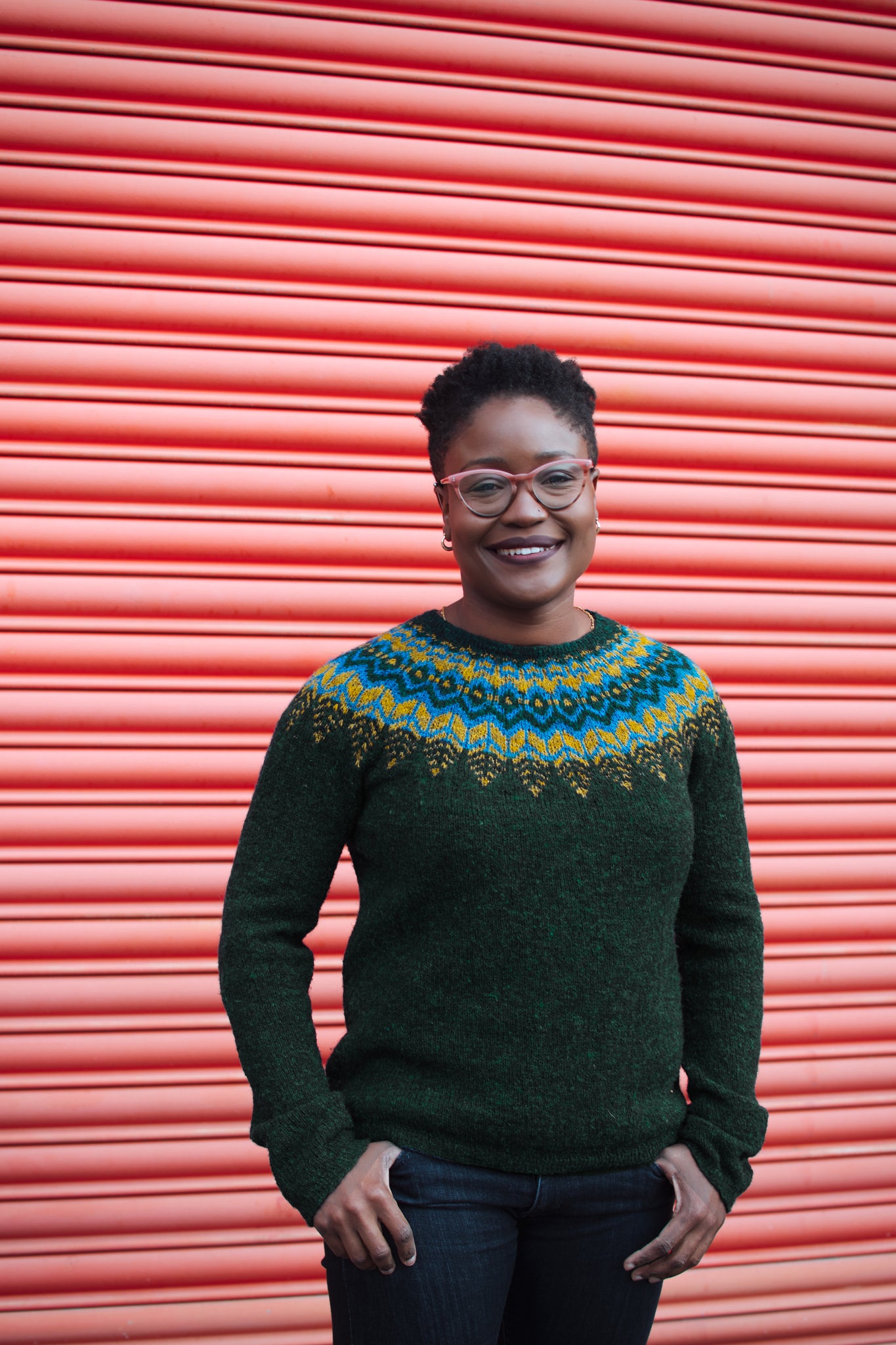 Black woman with short hair and glasses wearing a dark greenish black threipmuir with blue and yellowish green yoke