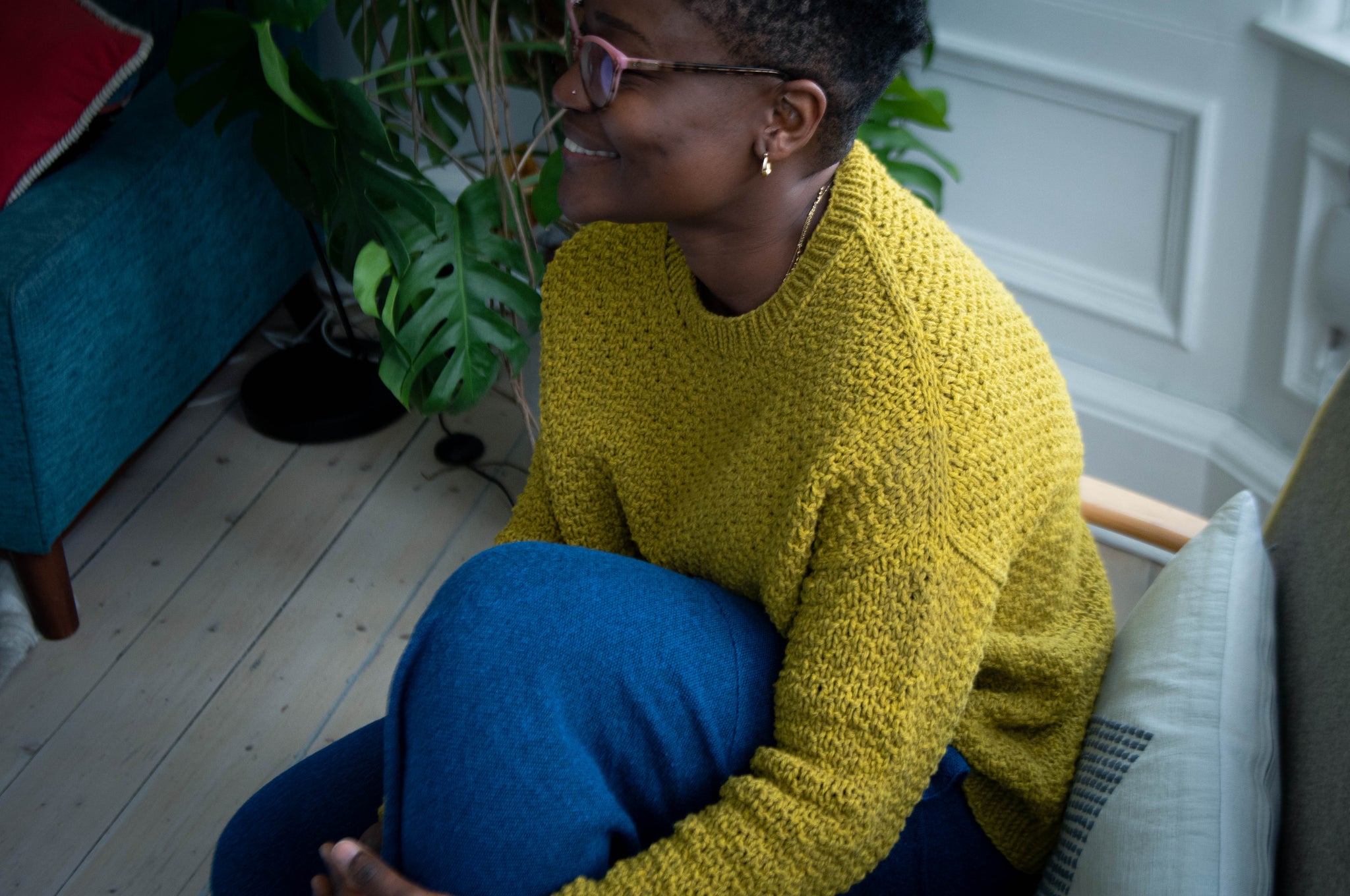 A black woman with short hair sits on a chair indoors with one knee held to her chest. She wears a yellow sweater with blue trousers and smiles while looking to the left of the image