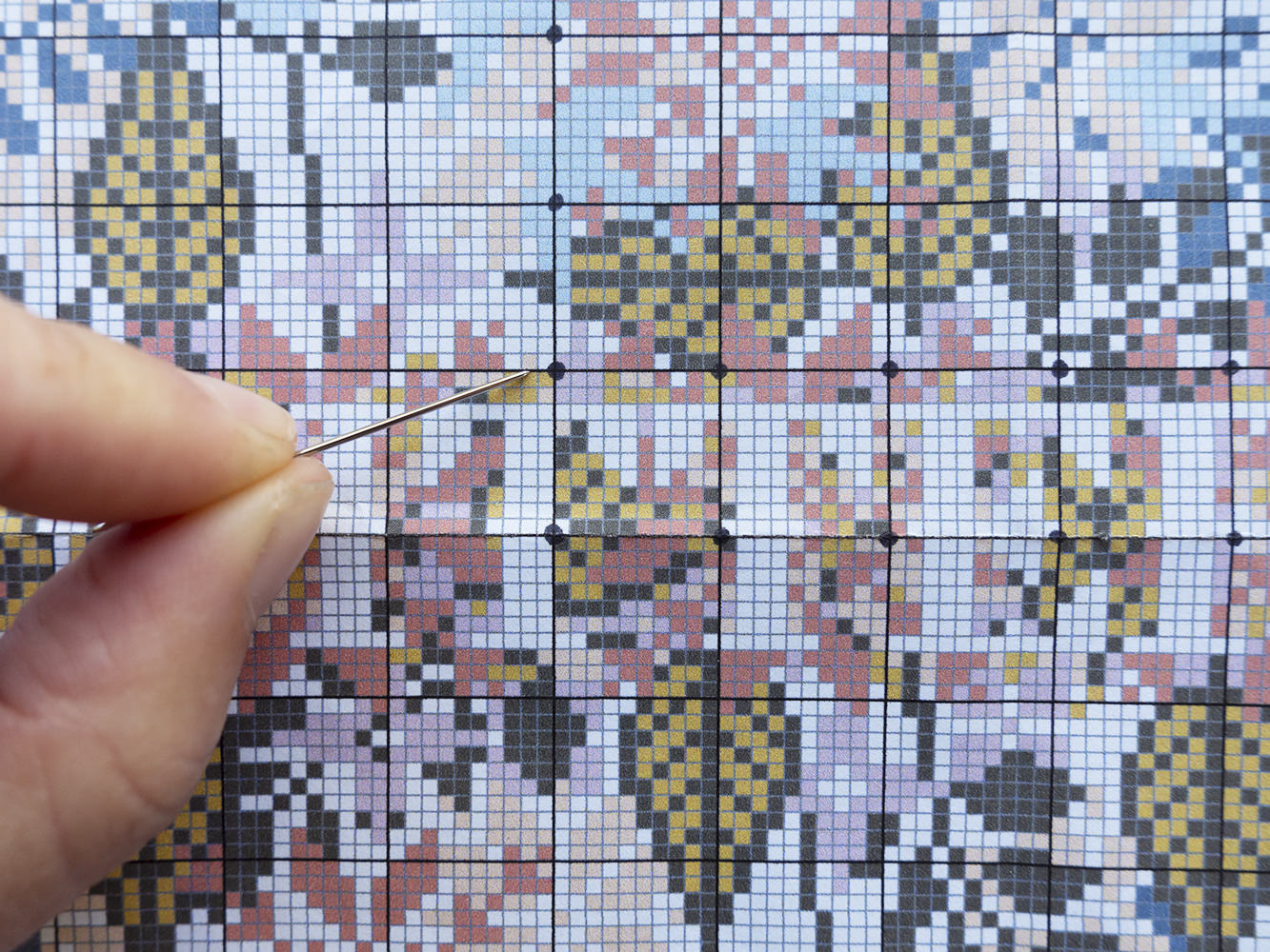 close up of knitter cross stitch chart with Zoe's hand holding a needle pointing at the centre gridline