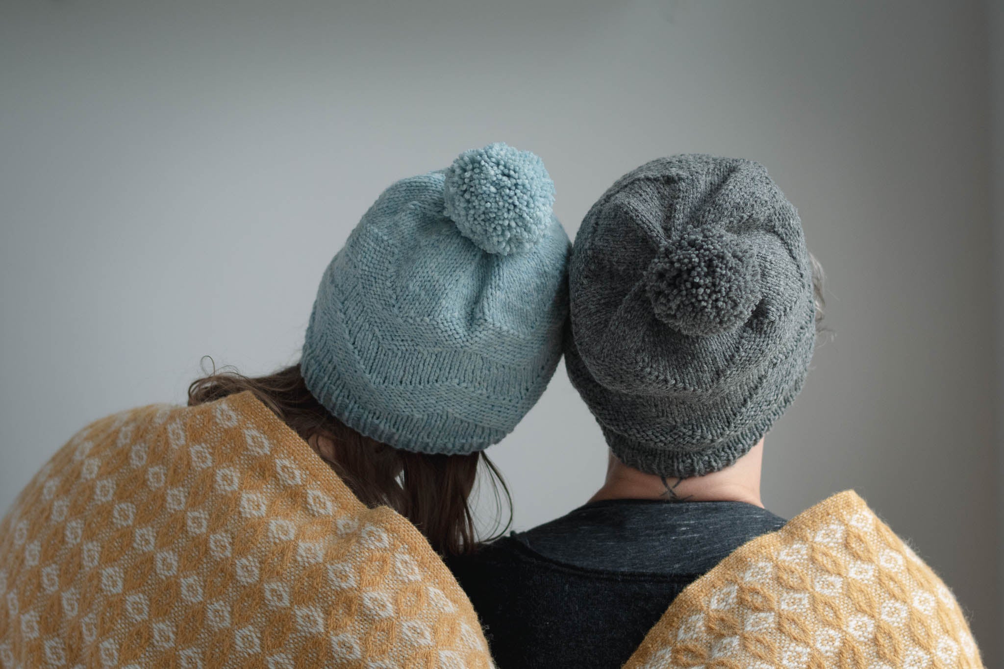 Two models face away from the camera, each wearing the same slouchy beanie hat with pom pom. They are snuggled together and wrapped in a shared yellow blanket.