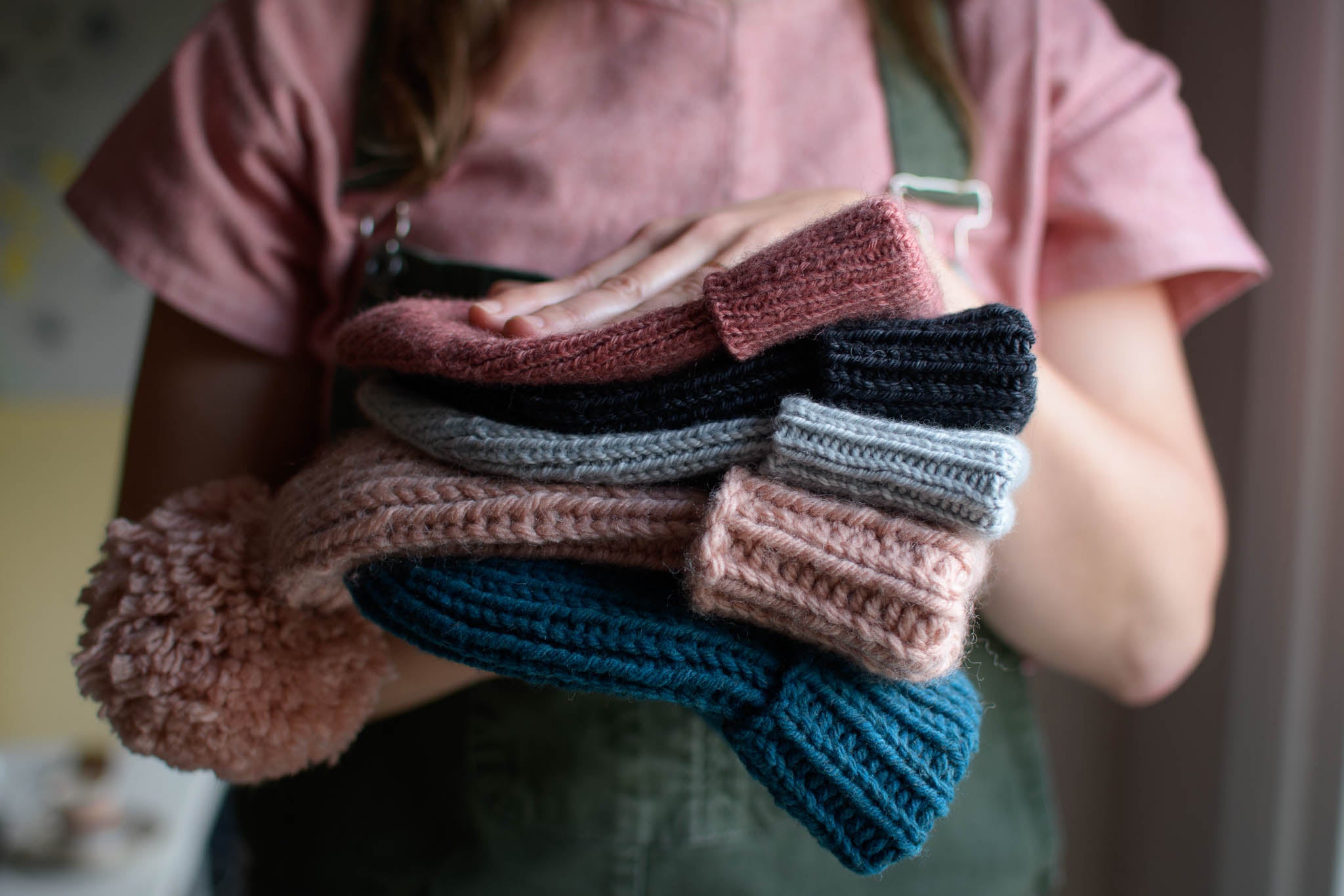 a person wearing a pink shirt and khaki dungarees holds a stack of ribbed hats in pink, pale blue, charcoal and teal