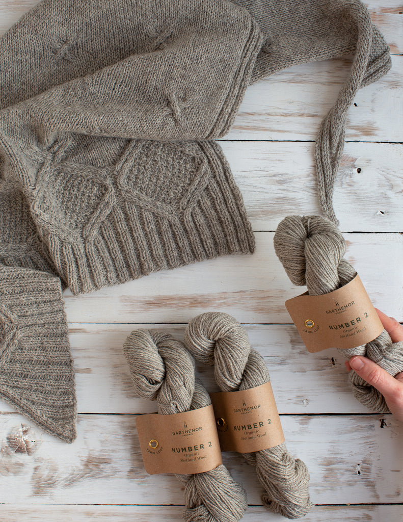 Flat lay photo with a cabled shawl draped in the upper left corner and 3 skeins of the same yarn in the bottom right corner. The yarn is a natural grey and there is a hand holding one of the skeins.