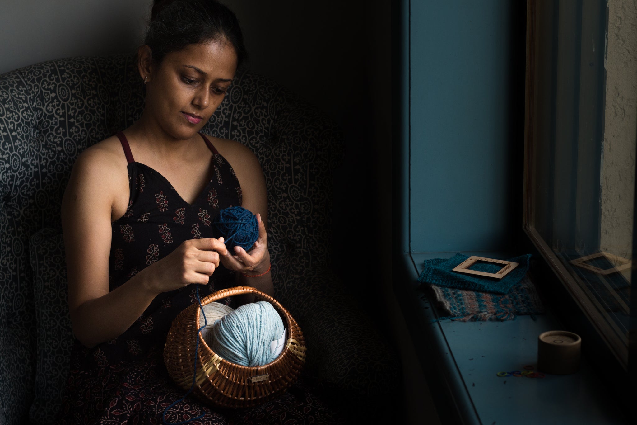 a women sits by a window against a dark wall, wearing a black dress. She is hand winding a ball of pale blue yarn, and there are swatches of knitting next to her on the windowsill.