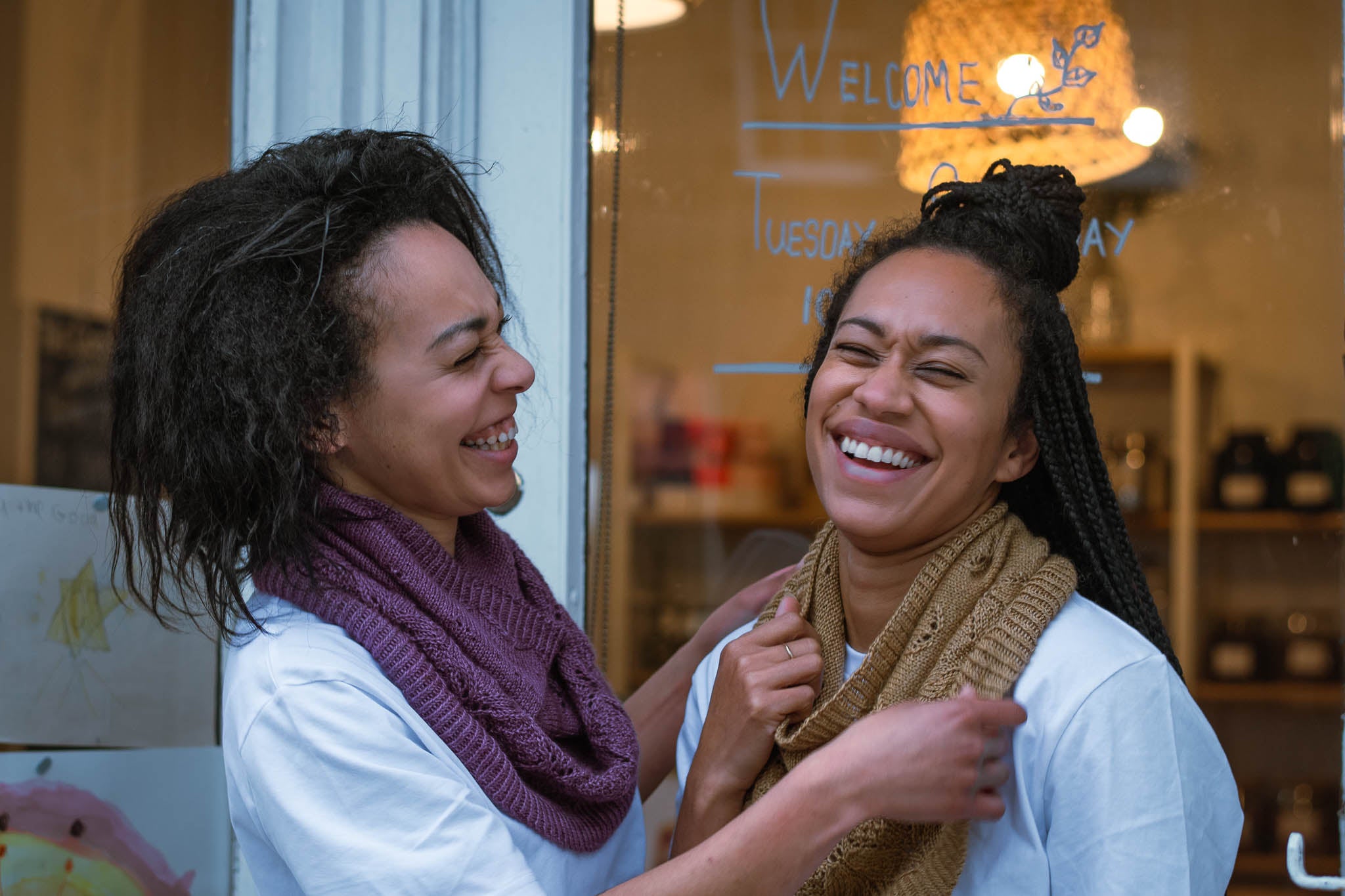 two Black women wearing white t shirts are pictured laughing together outside a shop, one is fixing the other's cowl. They're both wearing lace patterned cowls in a drapey yarn, one is purple and the other gold
