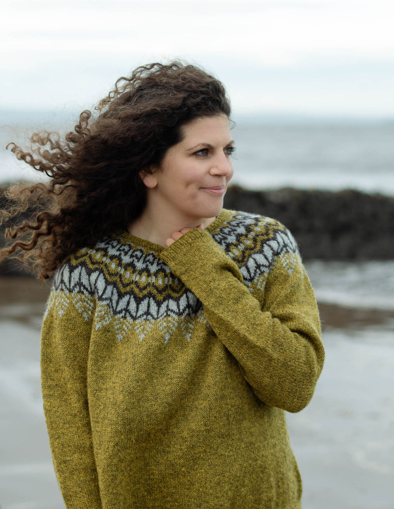 A white woman with long curly brunette hair stands on the beach smiling at the camera with the sea behind her. She is wearing an earthy green knit sweater with a dark brown and light grey colourwork yoke. Her left arm is folded up and her hand is near her neck holding the cuff of her sweater. 