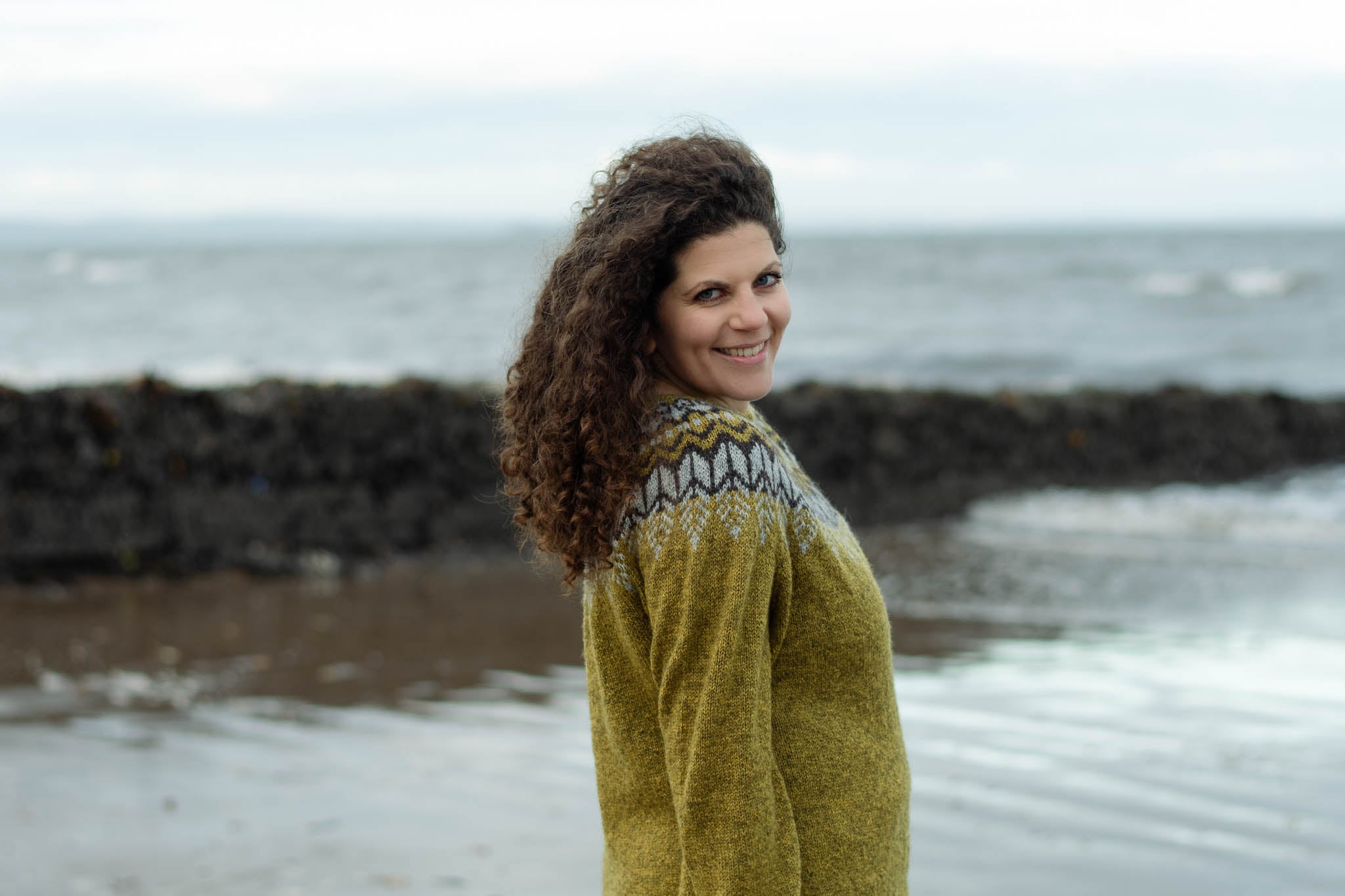 A white woman with long curly brunette hair stands on the beach smiling at the camera with the sea behind her. She is wearing an earthy green knit sweater with a dark brown and light grey colourwork yoke. Her arms hand down by her sides.
