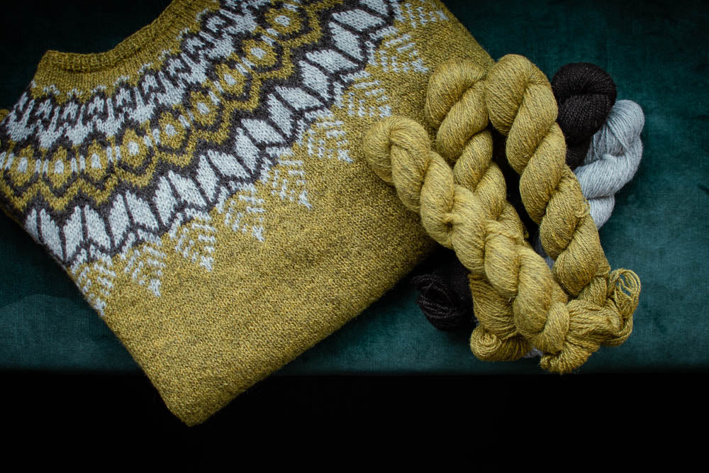 a gold green sweater with contrast yoke in grey and brown folded on a flat surface, with skeins of yarn overlapping at the side.