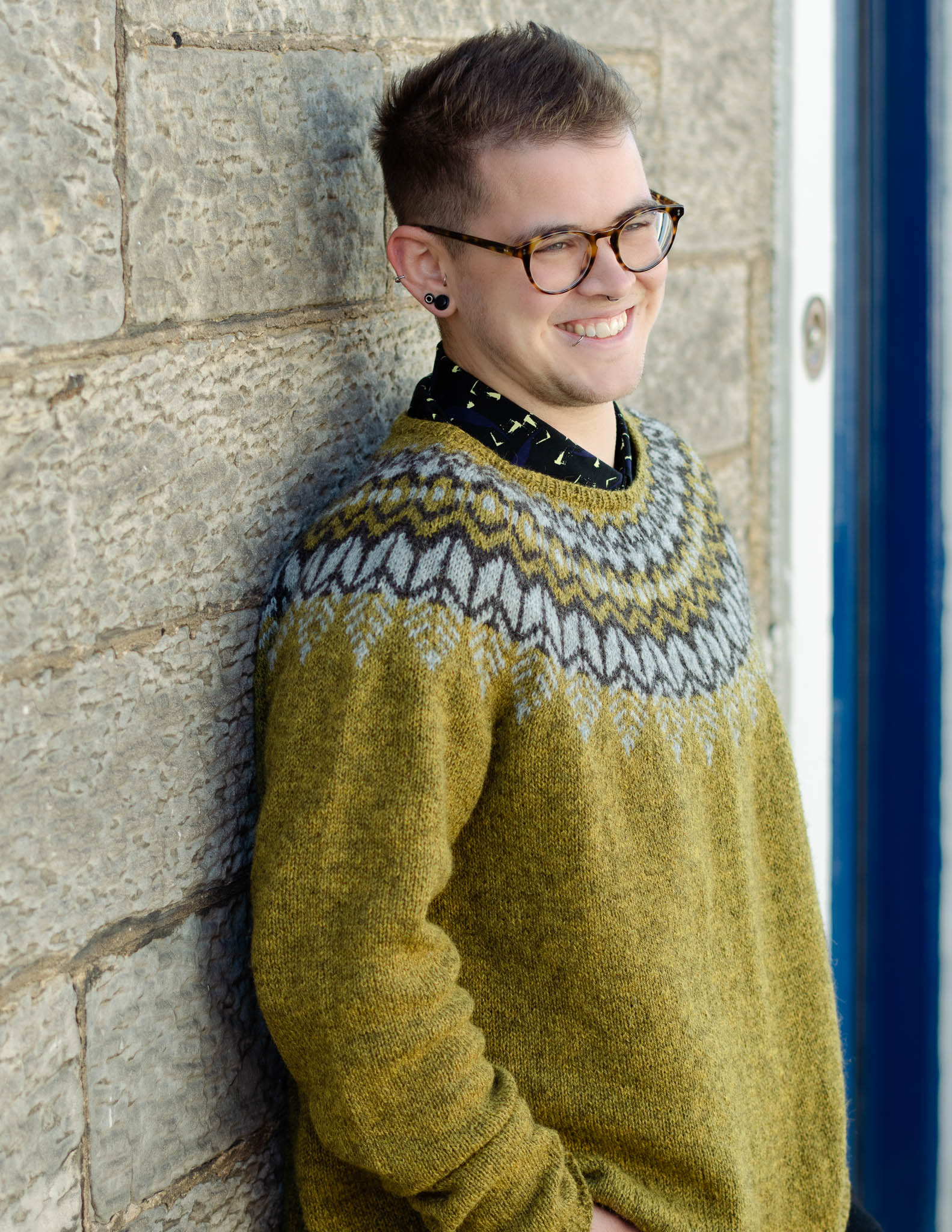 A white man leans casually against a light coloured stone wall, he has short hair, is wearing dark glasses and smiles at the camera. He is wearing an earthy green knit sweater with a dark brown and light grey colourwork yoke and dark trousers.