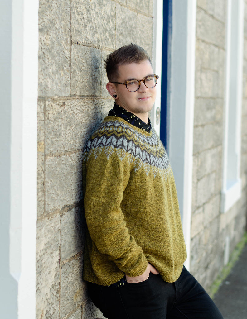 A white man leans casually against a light coloured stone wall, he has short hair, is wearing dark glasses and smiles at the camera. He is wearing an earthy green knit sweater with a dark brown and light grey colourwork yoke and dark trousers. His left hand is in his front pocket.