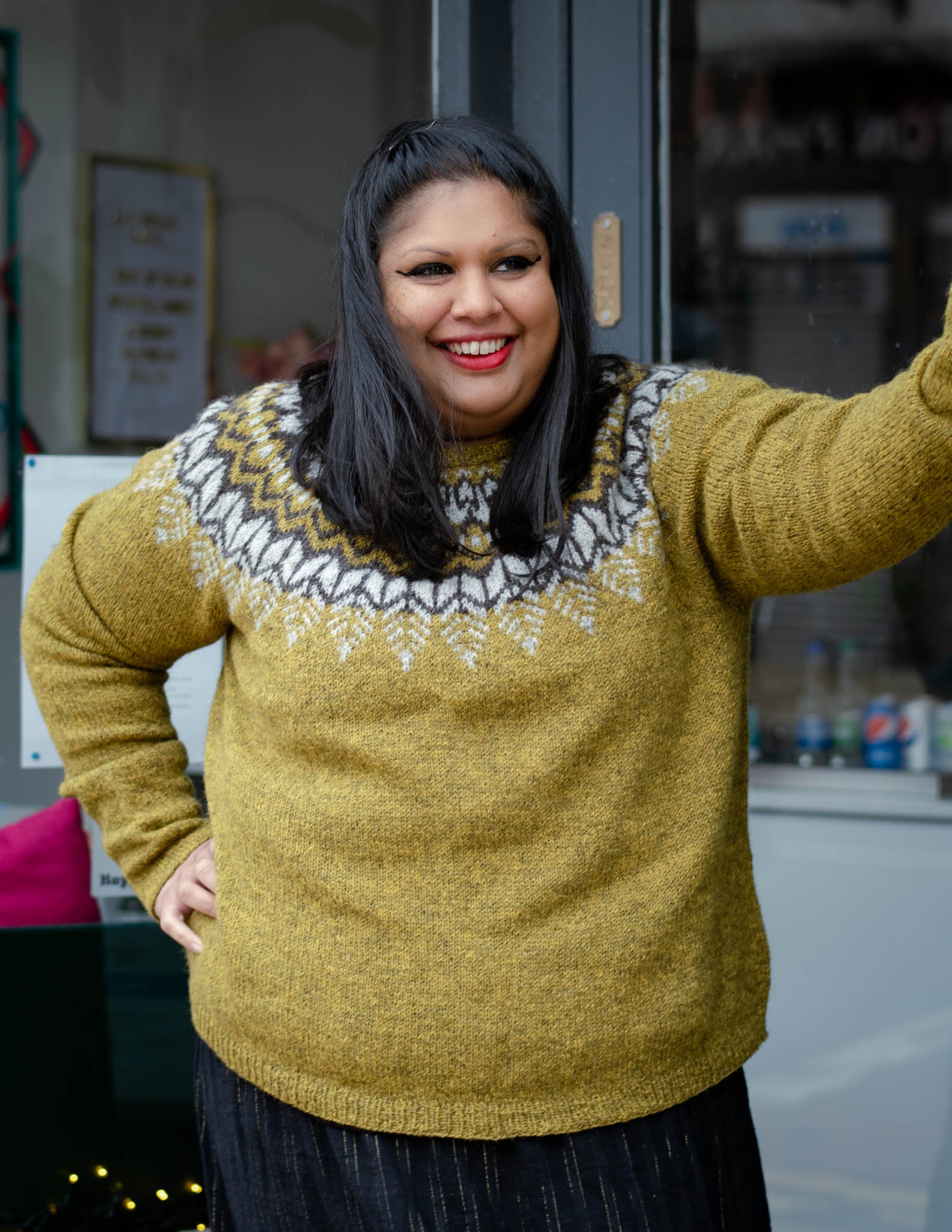 A south asian woman with mid length dark hair stands in a doorway looking towards the right of the frame. She is wearing  an earthy green knit sweater with a dark brown and light grey colourwork yoke.