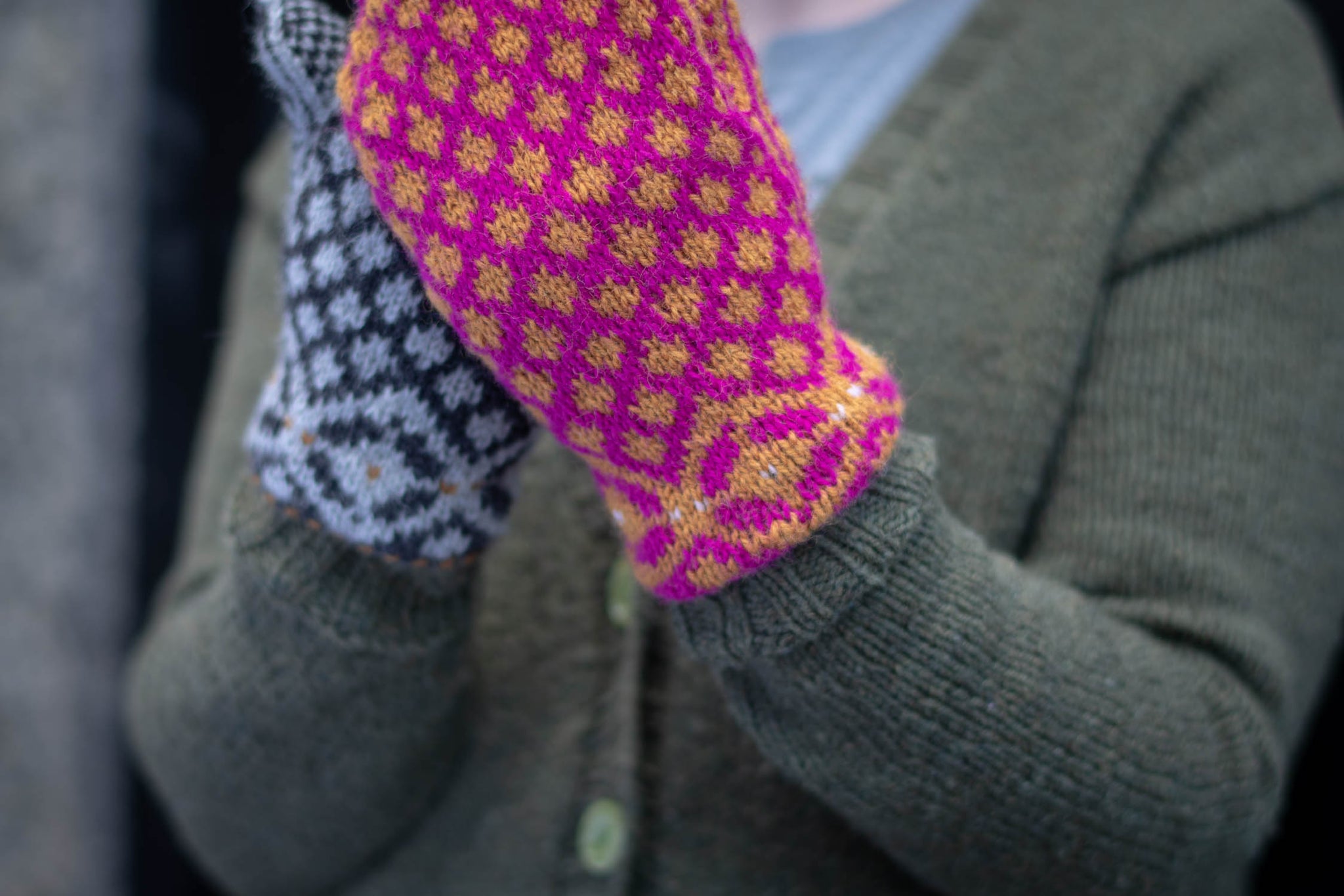 Two hands held up, clasped together. One is wearing a pink and yellow colourwork mitten, the other wears a grey mitten.