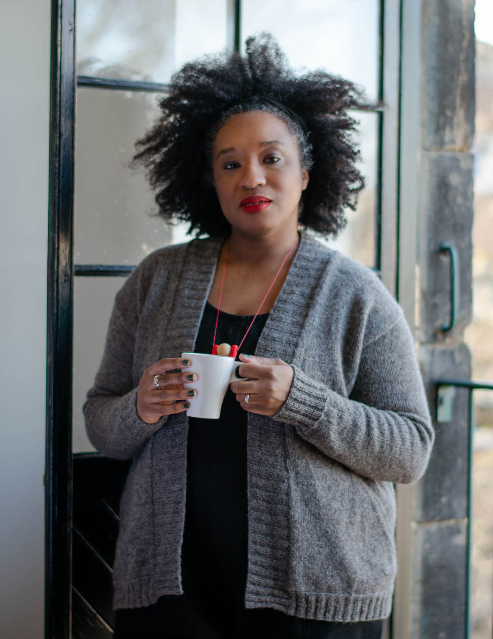 A black woman with an afro, stand in front of an open balcony door holding a white coffee mug. She's wearing a grey cardigan with a wide ribbed band, and pockets over a black jumpsuit.