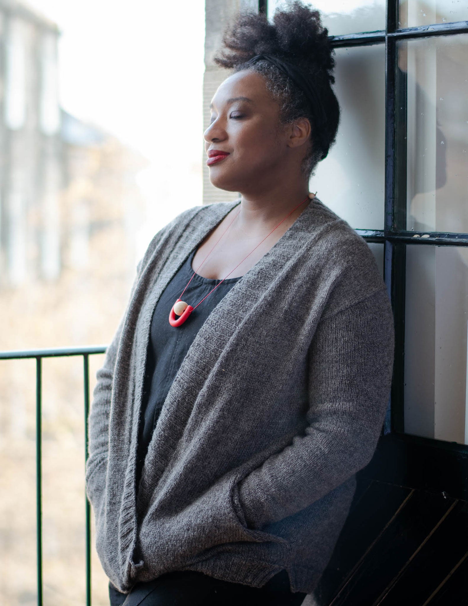 A black woman with her hair tied up on top of her head, leans against and open balcony door she got her eyes closed and looks blissful, her hands are in the cardigan pockets. She's wearing a grey cardigan with a wide ribbed band, and pockets over a black jumpsuit.