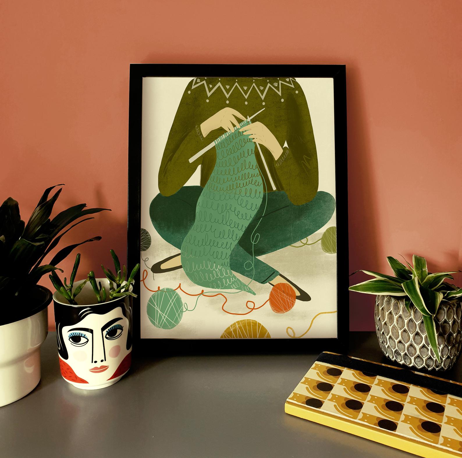 A print of a knitter from the shoulders down sitting cross legged and knitting a green scarf on straight needles. Next to the framed print are houseplant in pots.