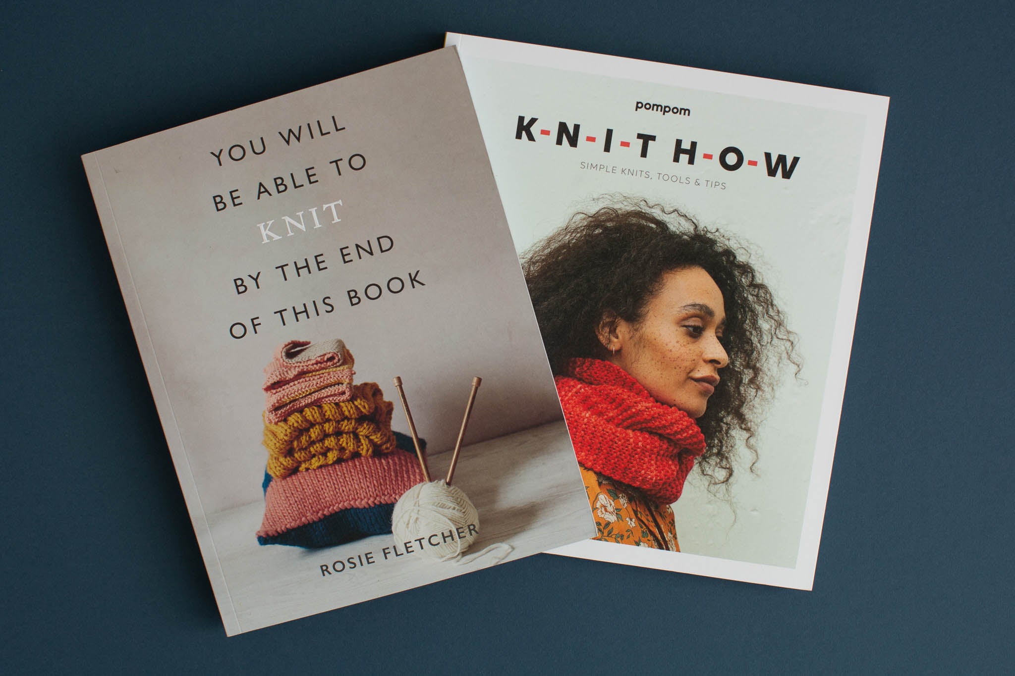 Dos libros colocados en horizontal, ligeramente superpuestos. Uno muestra una pila de tejidos y se titula 'You Will Be Ape to Knit By The End of This Book' y el otro muestra a una joven negra con un capó rojo llamado 'Knit How.''You Will Be Able To Knit By The End Of This Book' and the other shows a young black woman wearing a red cowl called 'Knit How.'