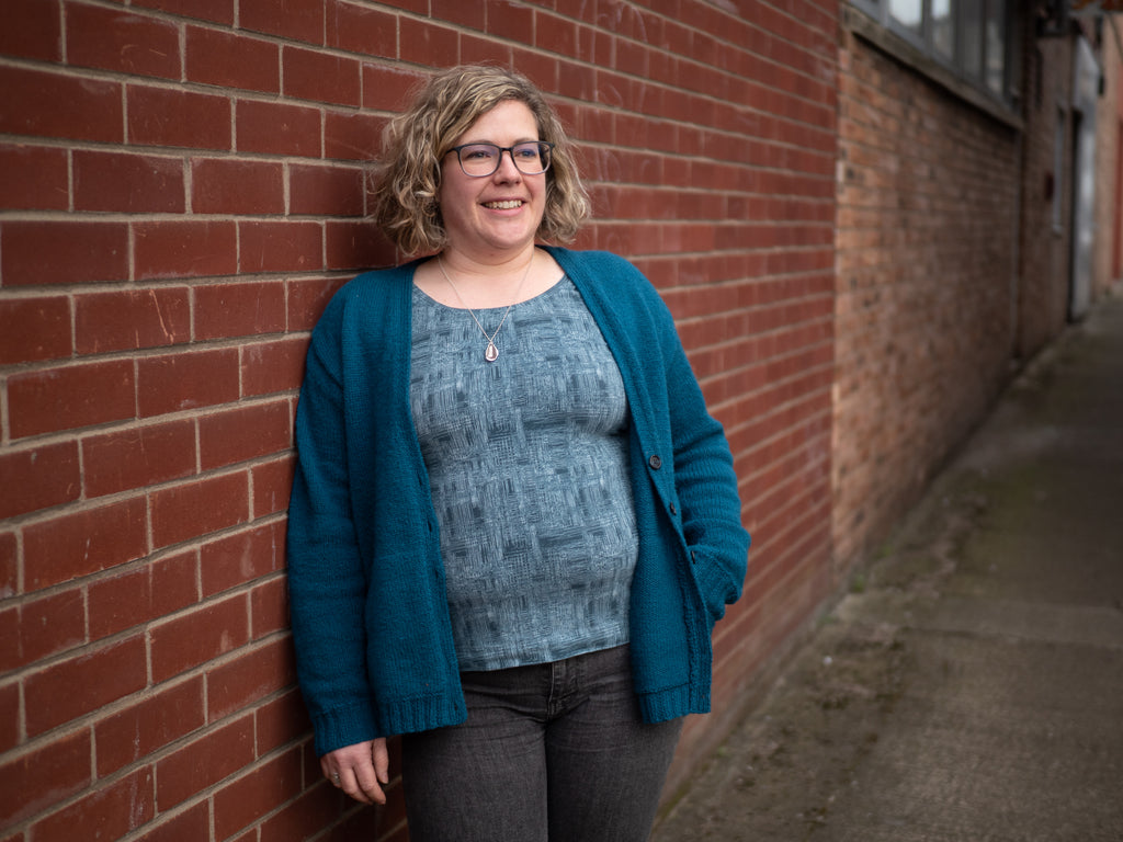 Woman with pale skin and brown curly hair leaning on a brick wall wearing a pale blue patterned top with the blue wardie, it fits closely