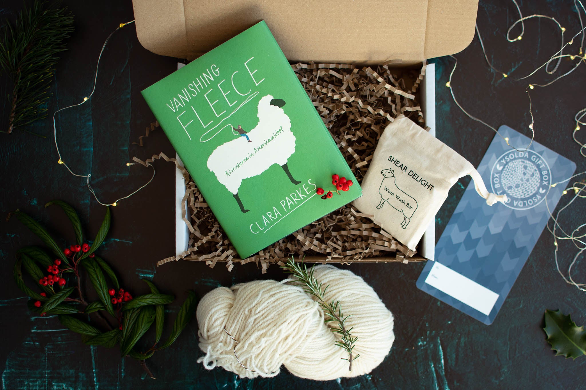 An image taken from above of an open box, containing a skein of white wool, a green book with a white sheep drawing on the cover, a bar of wool soap in a cotton drawstring bag.