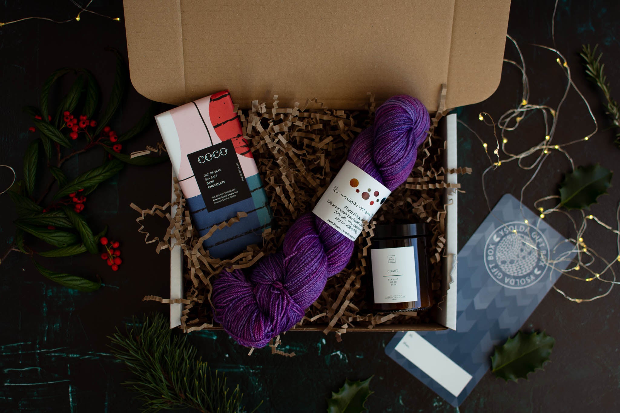 An open box containing a bar of chocolate, a purple skein of yarn and a candle.
