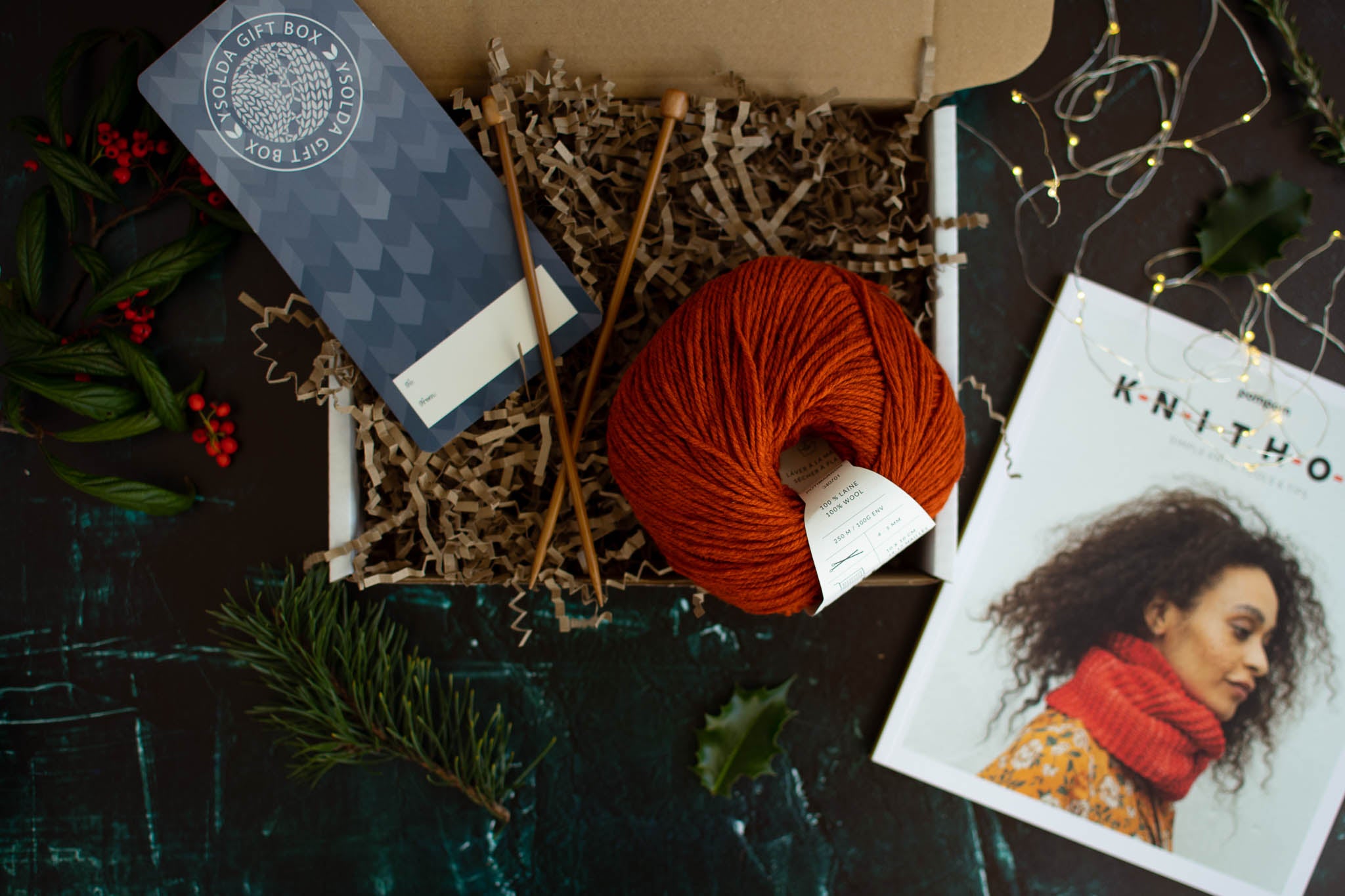An open box containing a pair of bamboo knitting needles, a red ball of wool and book called Knit How with a black woman wearing a red cowl on the cover.