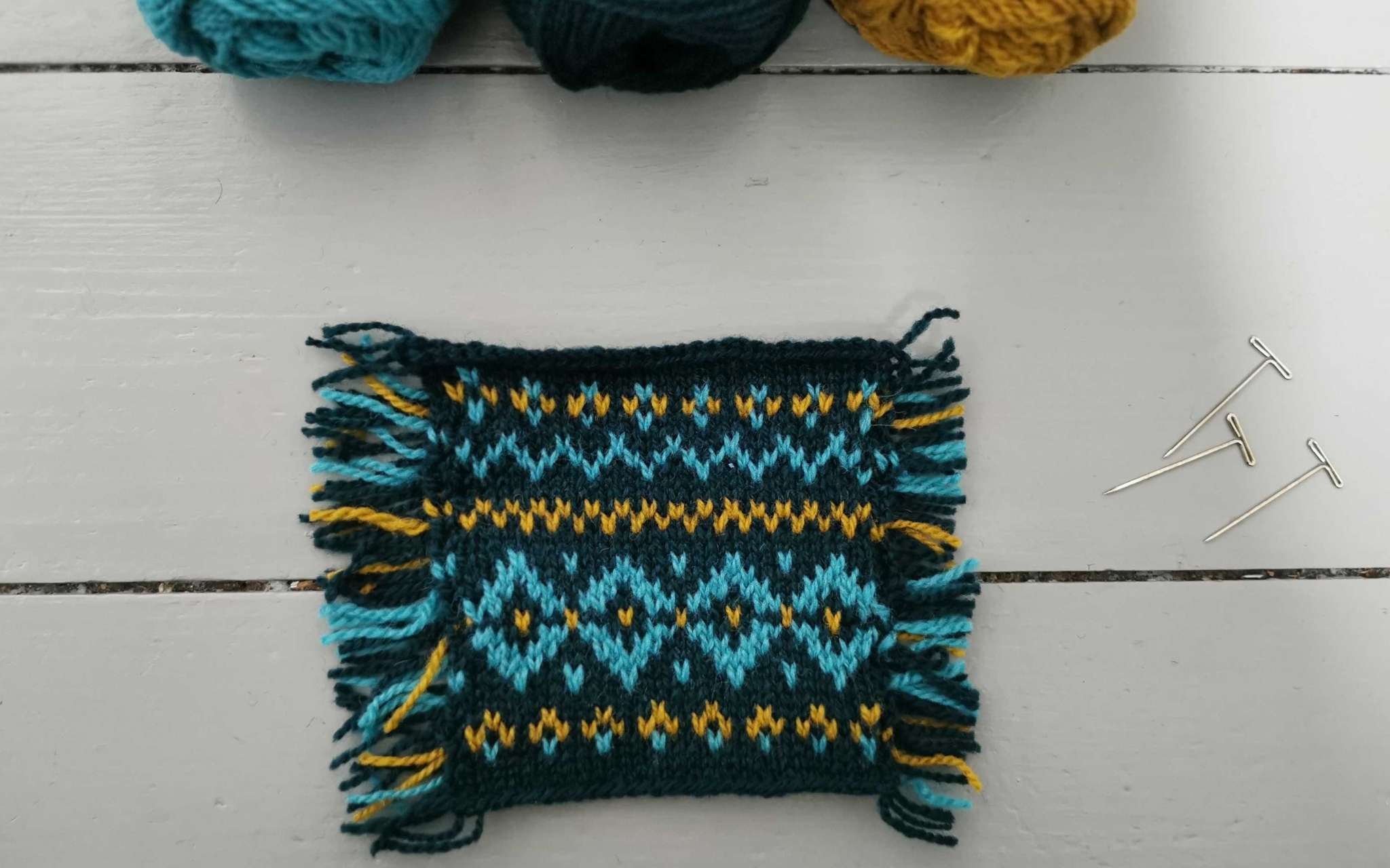 a square knitted swatch in navy, gold and teal colourwork. Three balls of yarn peak out at the top of the image and lay on a grey wooden surface