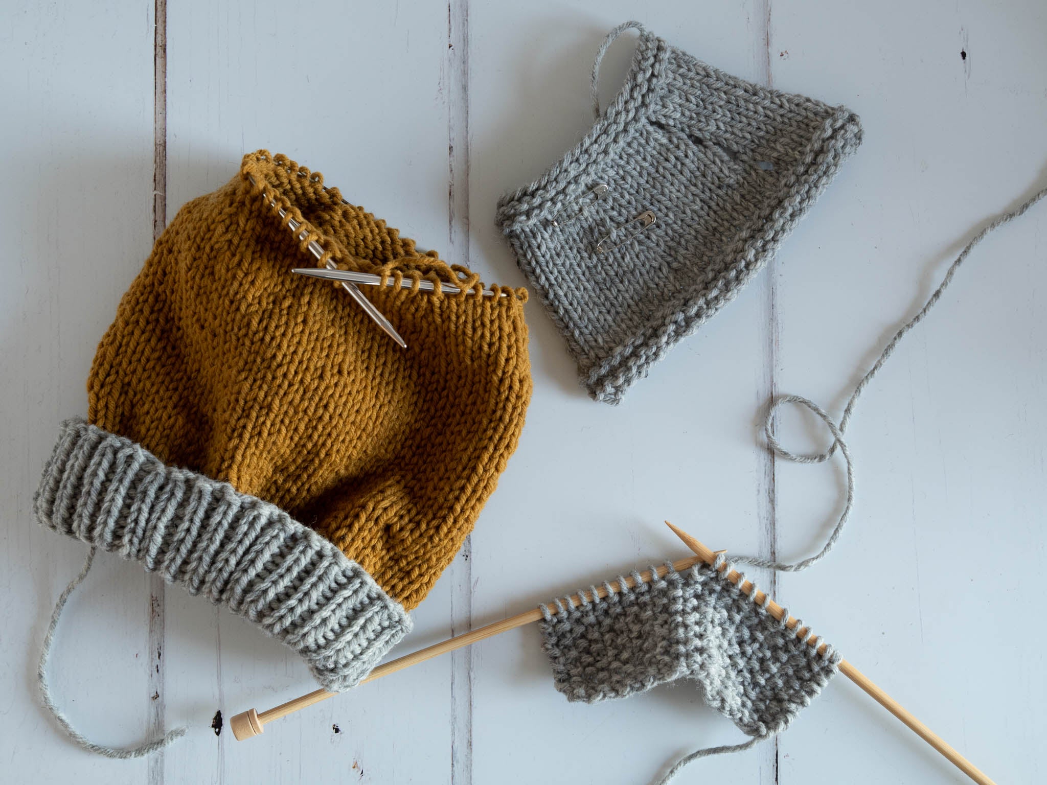 three pieces of knitting laying on a flat surface, two of which are grey swatches and one is a project in the round with a grey ribbed brim.