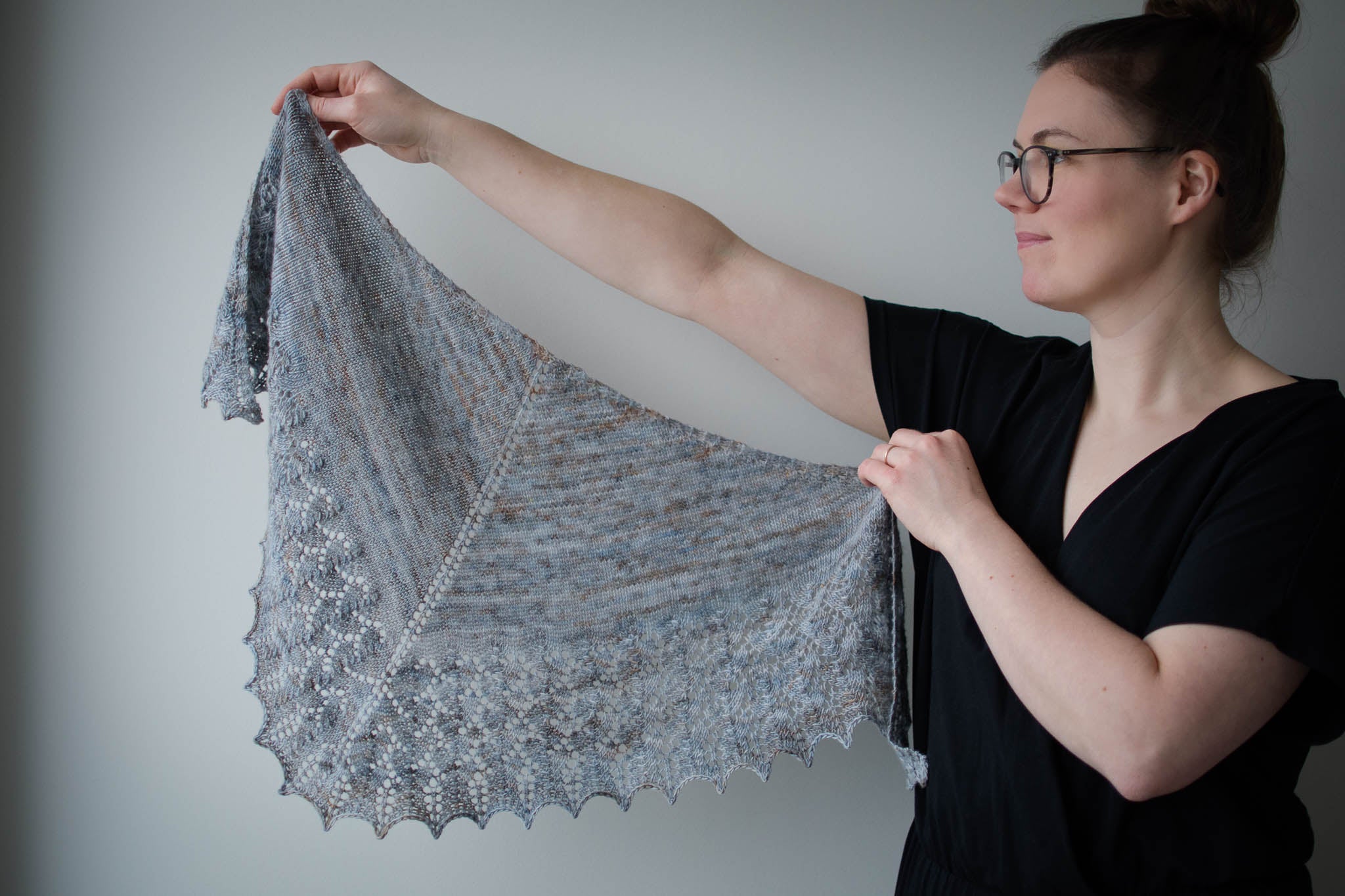 A young white woman wearing a black t-shirt holds up a grey lace shawl, spreading it out to the left side of the image.