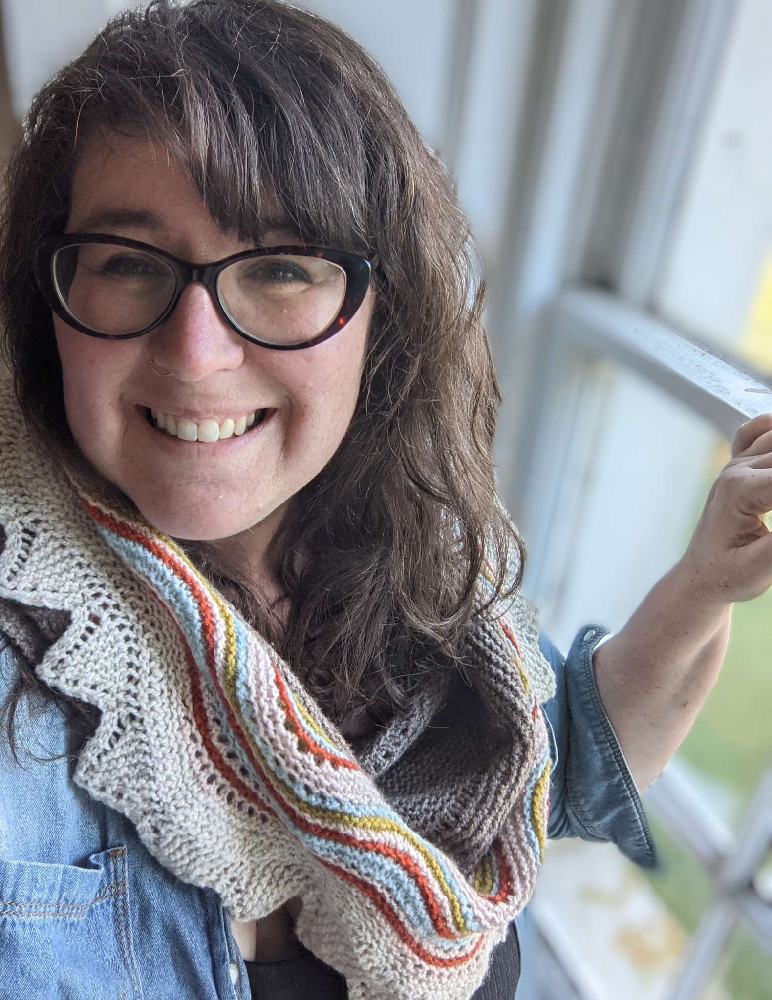 A white woman with long wavy hair and glasses stands indoors in front of a window. She is smiling and wears a neutral shawl with brightly coloured stripes and a blue shirt.