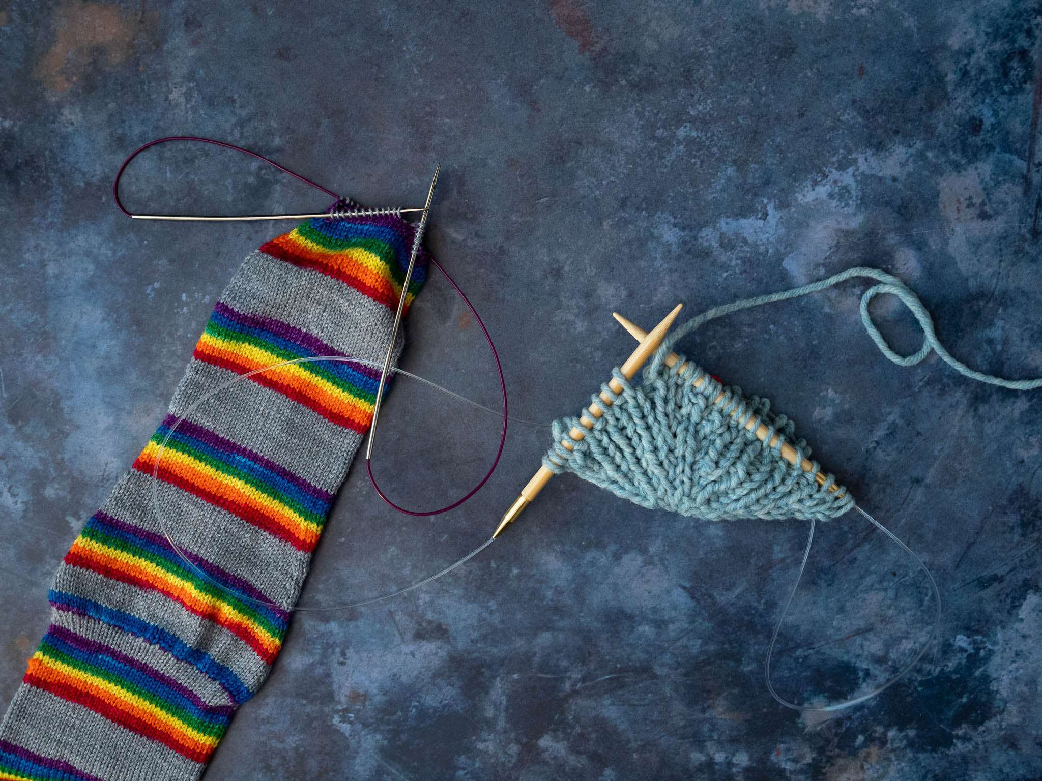 a rainbow striped sock and the beginning of a top down blue hat on long circular needles. Loops of cable can be seen on either side of each project