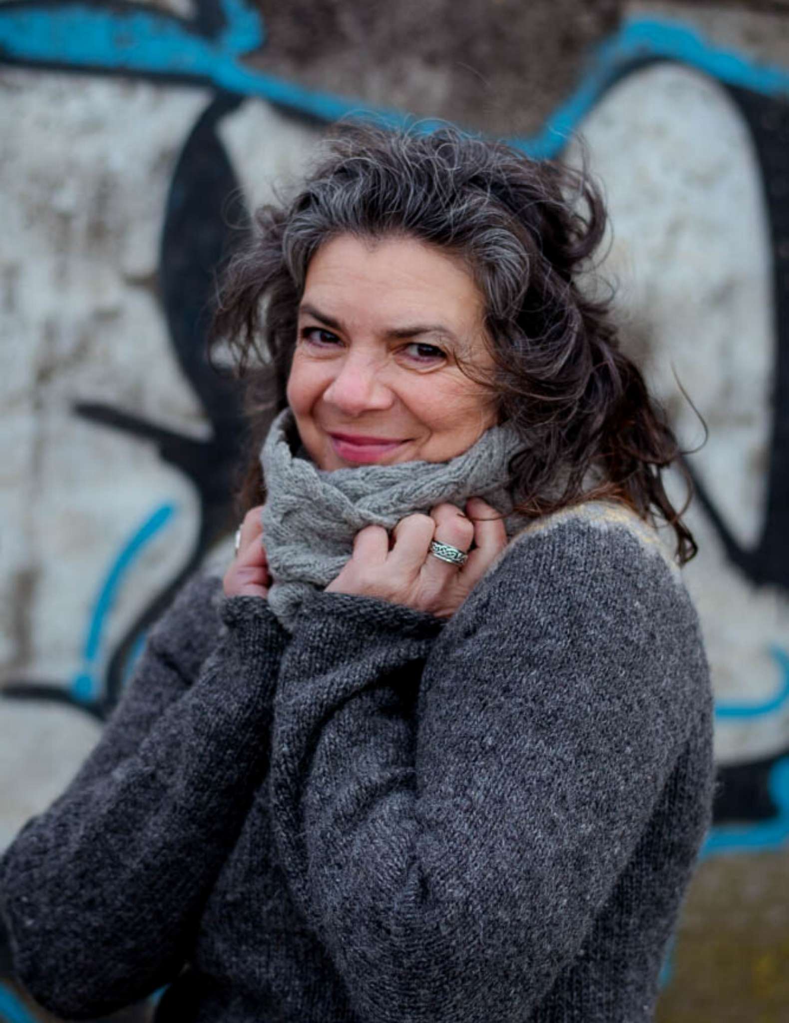 a white woman in her 50s with dark curly hair wears a grey sweater and pale grey cowl. Their hands are held up to the cowl and they are smiling.