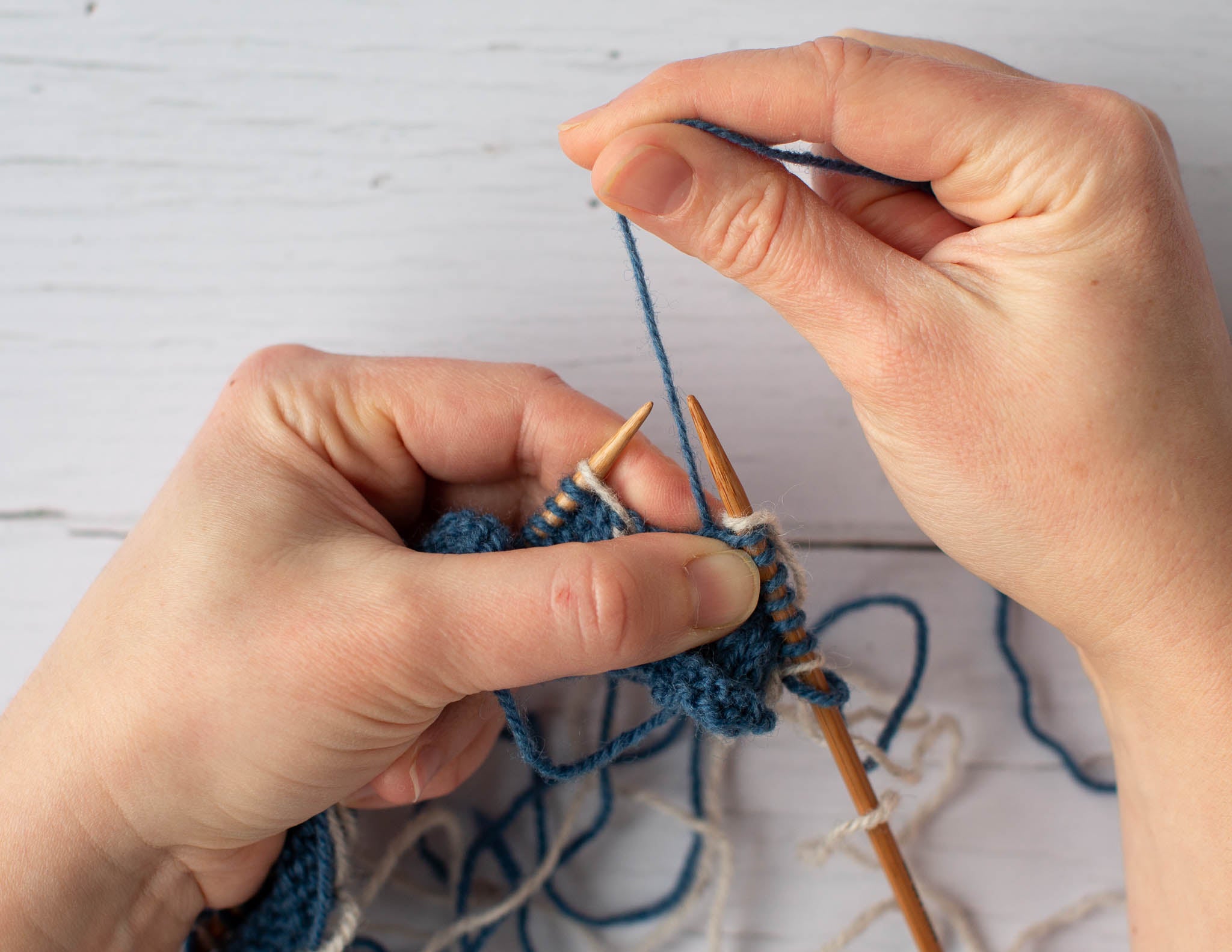 Two white hands holding a piece of blue and white knitting over a flat surface. The knitting is on two wooden needles and the right hand holds a strand of blue yarn high above the knitting in the centre.