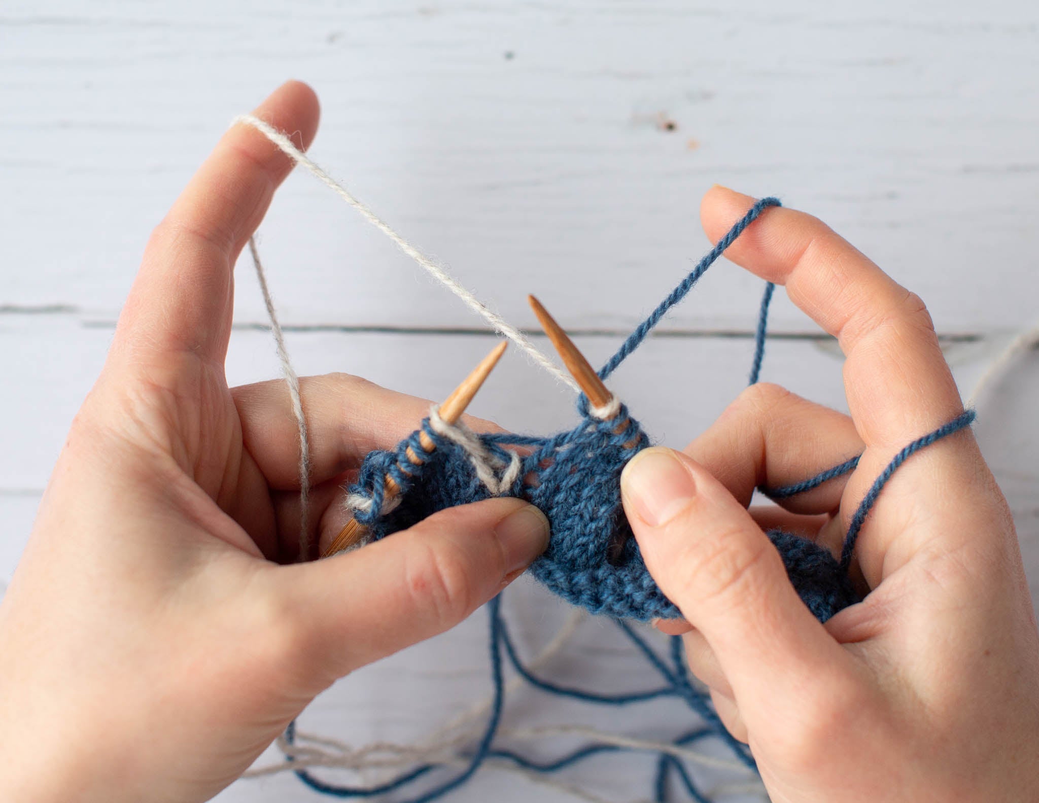 Two white hands hold a piece of blue and white knitting over a flat surface. The left hand has a strand of white yarn wrapped around the index finger, and the hand holds the blue yarn.
