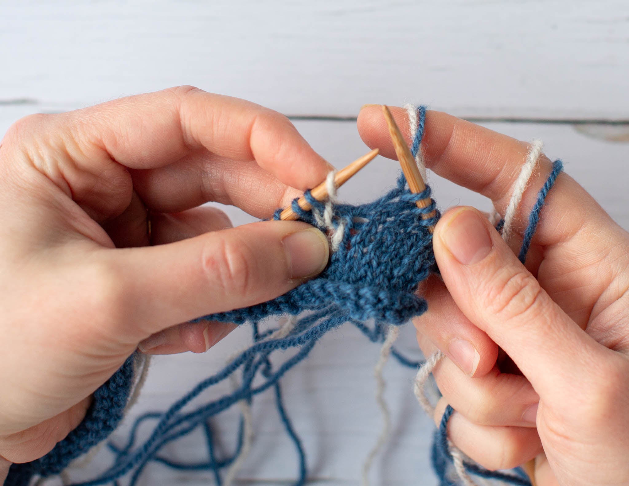 Two white hands hold a blue and white piece of knitting, on wooden knitting needles. The left hand grips the fabric and the right hand has a blue and white strands of yarn wrapped twice around the index finger.