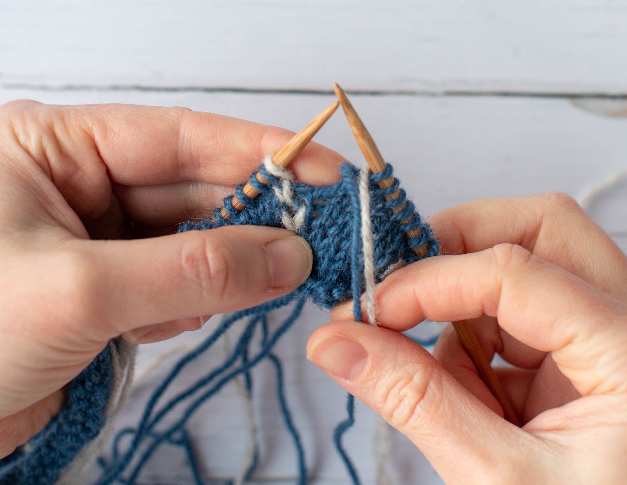 Two white hands hold a blue and white piece of knitting over a flat surface, the left hand grips the fabric and the right thumb and index finger hold a blue and white strand of yarn downwards.