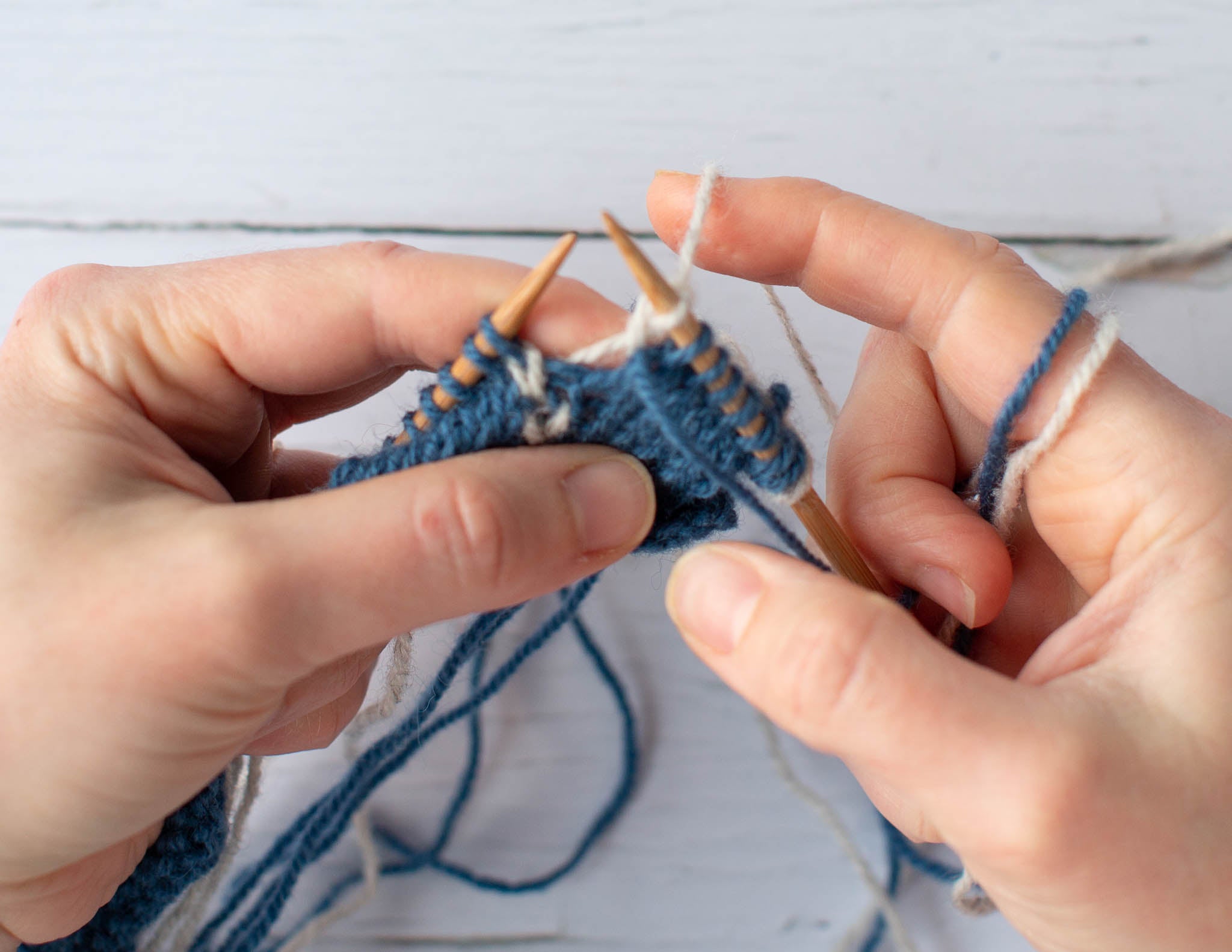 Two white hands holding a blue and white piece of knitting on wooden needles.
