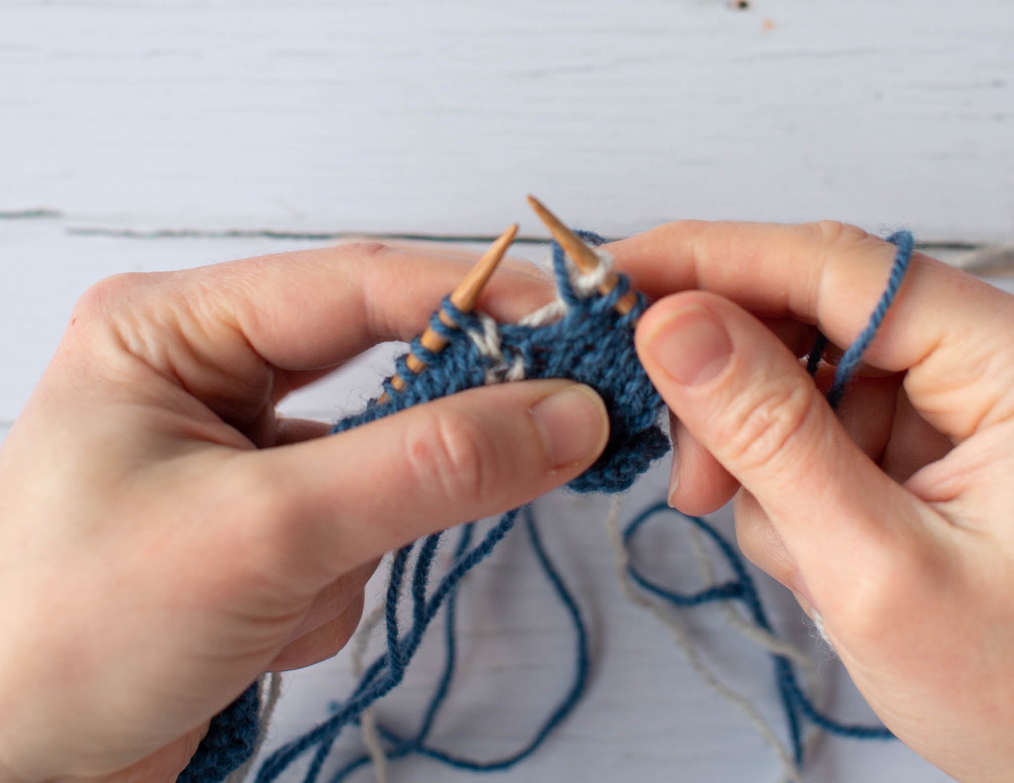 Two white hands holding a blue and white piece of knitting on wooden needles.