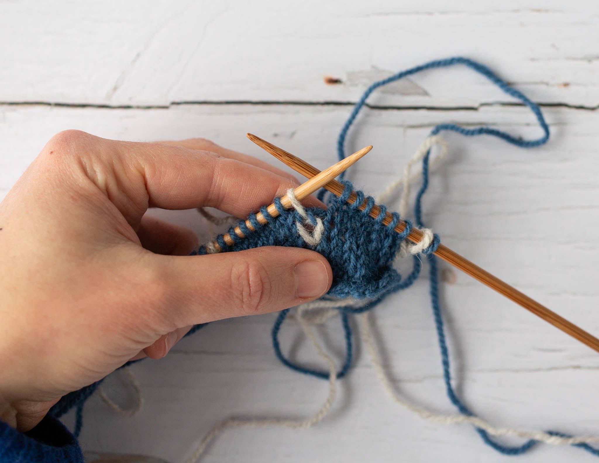 a white hand in the left of the image holds a small piece of blue and white knitting, still on wooden needles with strands of working yarn looped around behind on a flat surface