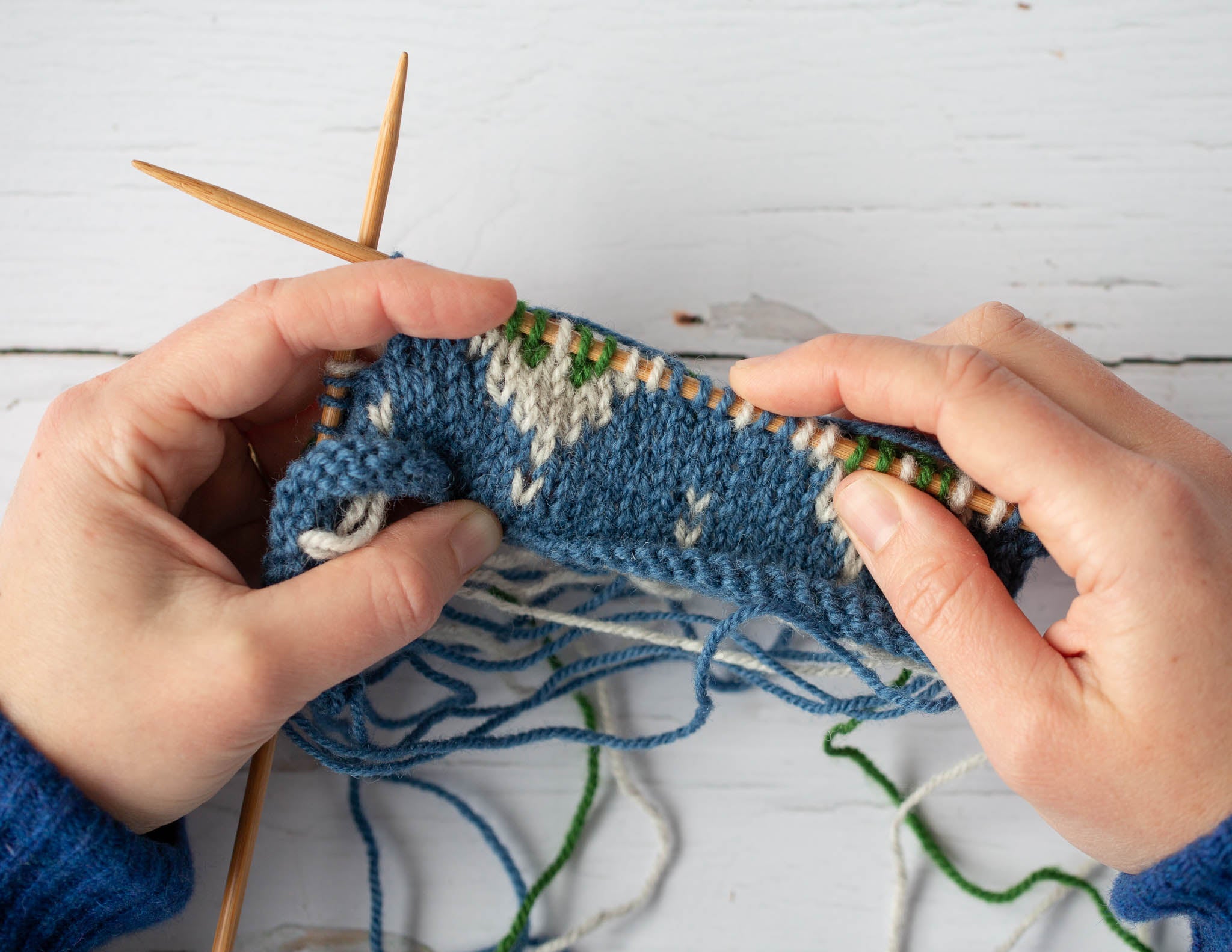 Two white hands hold a piece of blue and white knitting over a flat surface. The fabric is still on wooden knitting needles and shows a colourwork pattern.