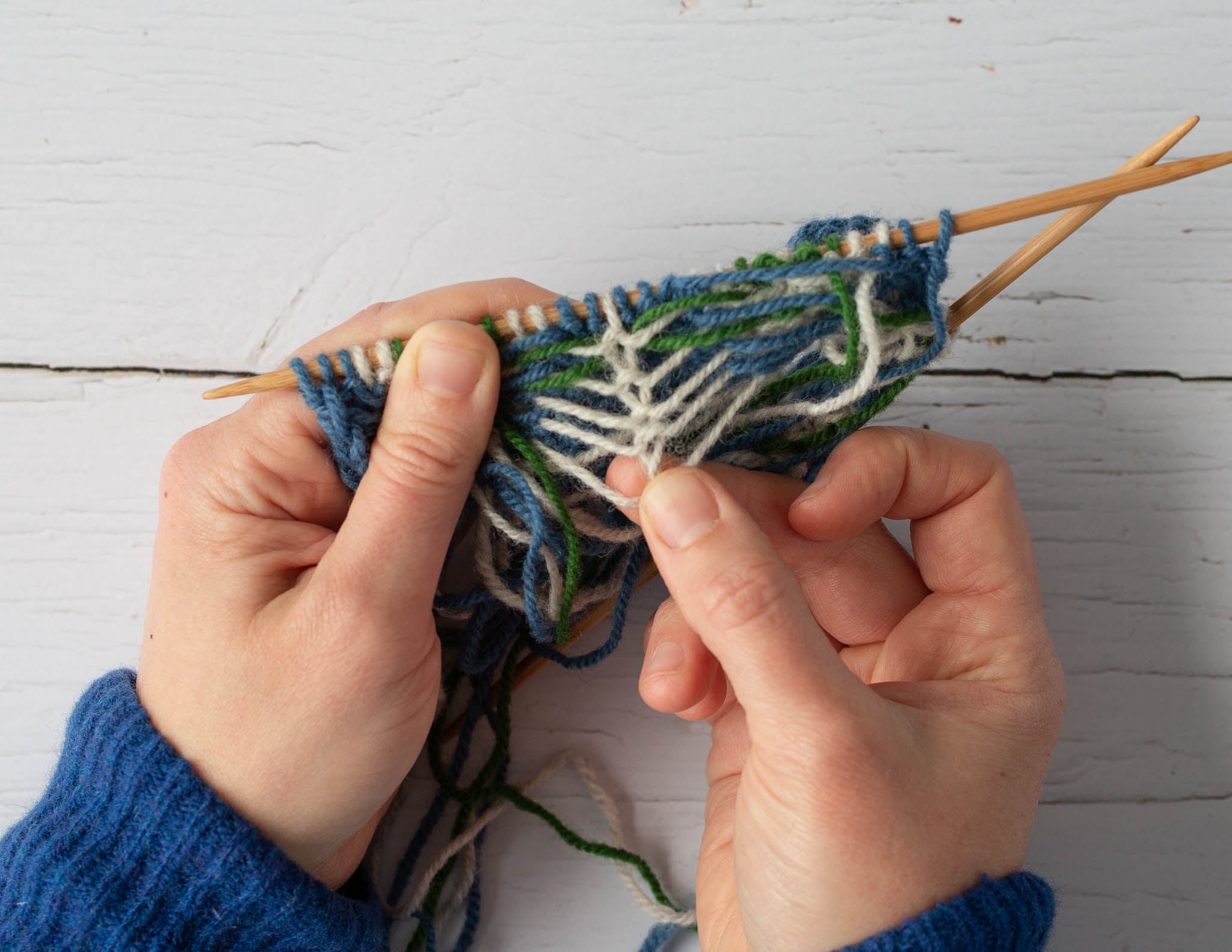 Two white hands hold a piece of blue and white knitting over a pale flat surface. The left hand grips the knitting near the knitting needles and the right hand pulls down on the fabric, showing the ladderback.