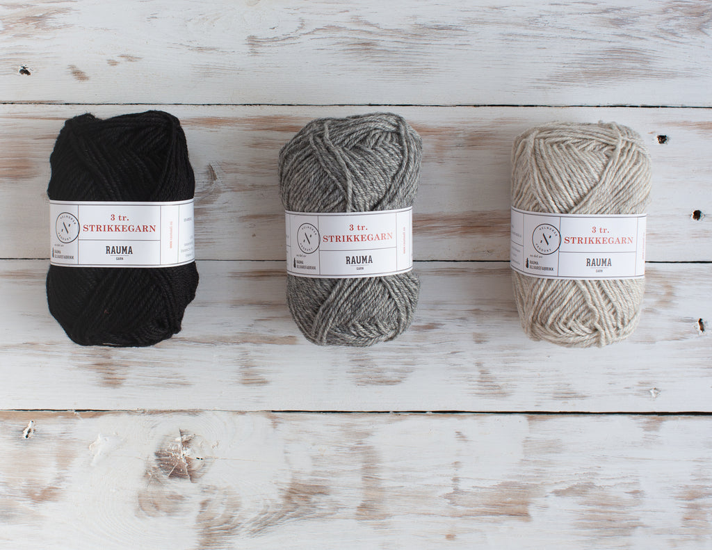 Three balls of Strikke yarn in black, mid grey and light grey. On a white-washed wooden background.