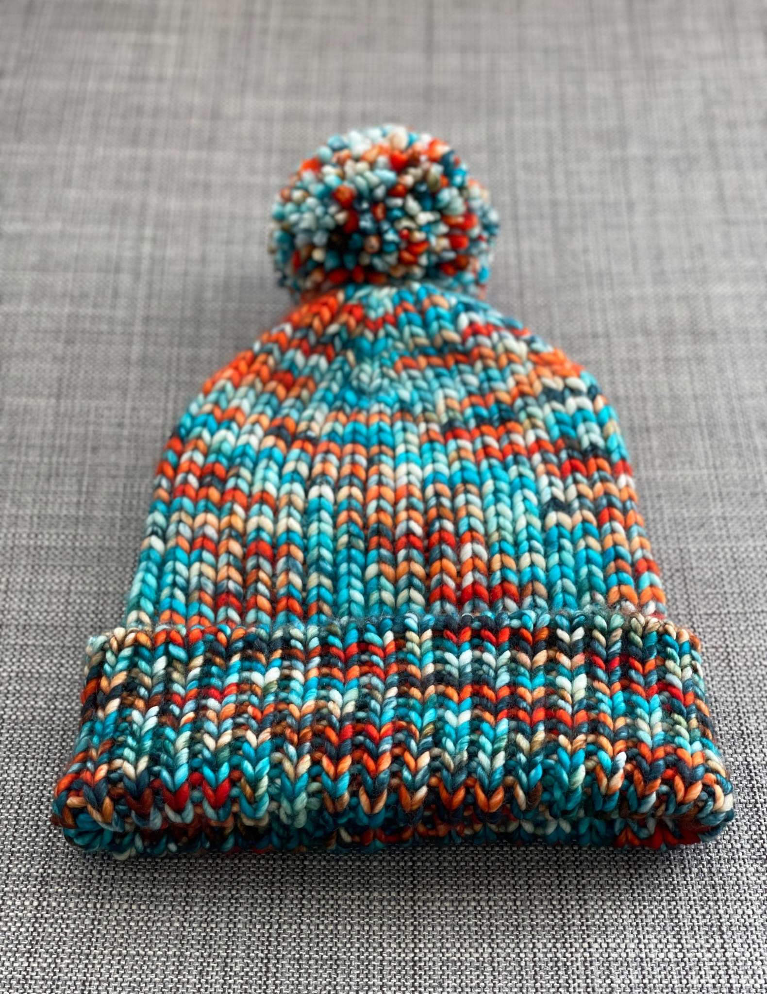 A variegated orange and turquoise ribbed hat with a fluffy pom pom lies on a grey fabric background. The colours on the hat have pooled into wide diagonal stripes.