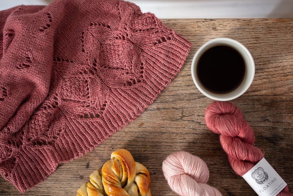 Reddish pink lace cowl draped in the corner of a wooden table next to 2 skeins of Floating yarn in the same colour and a paler version, a cup of coffee and a pastry.