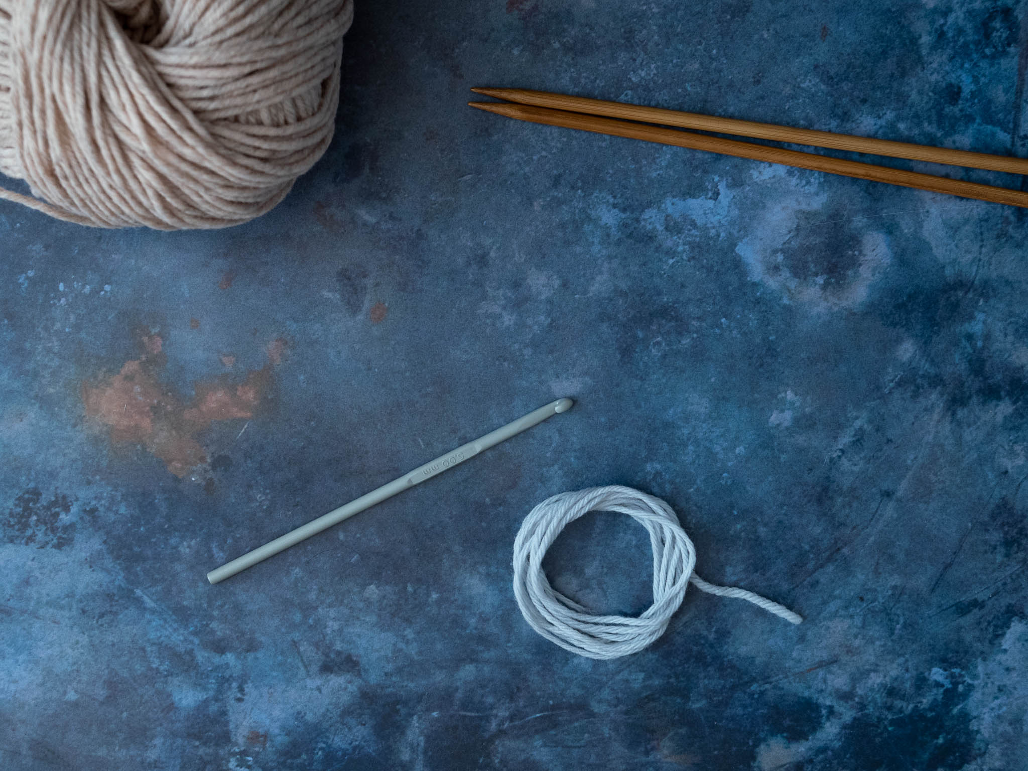 A mottled blue desk surface with a ball of beige yarn, a pair of knitting needles, some light blue scrap yarn, and a crochet hook.