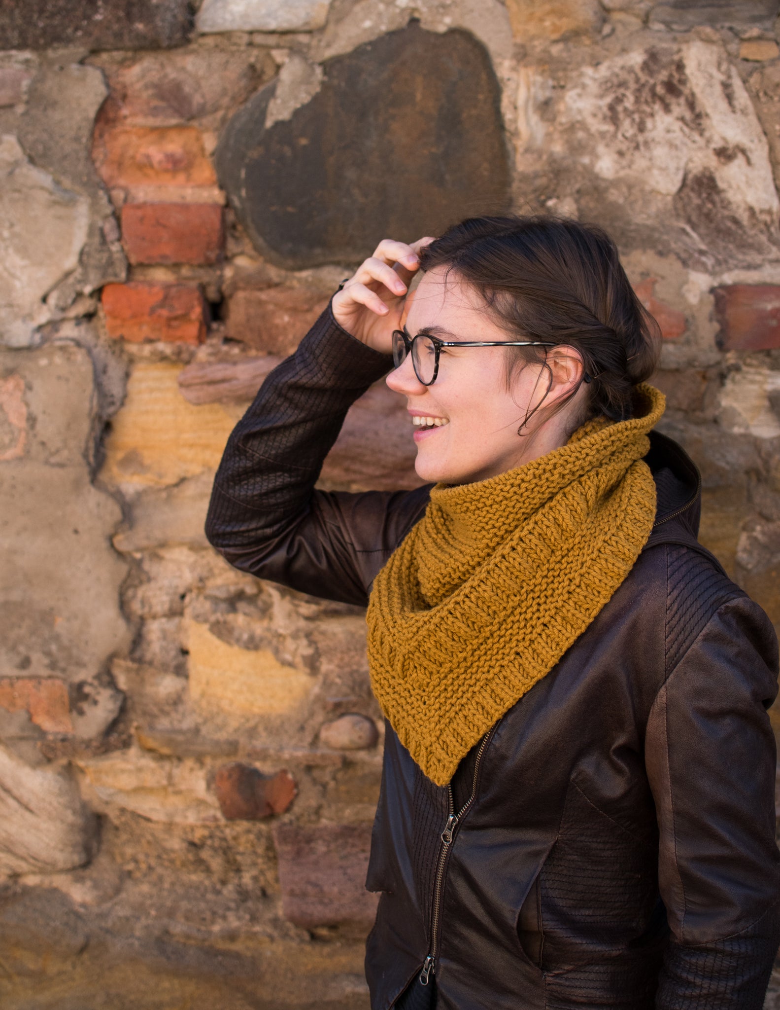 Picture of a white skinned woman wearing a gold bulky cowl. She is facing left against a brick wall.