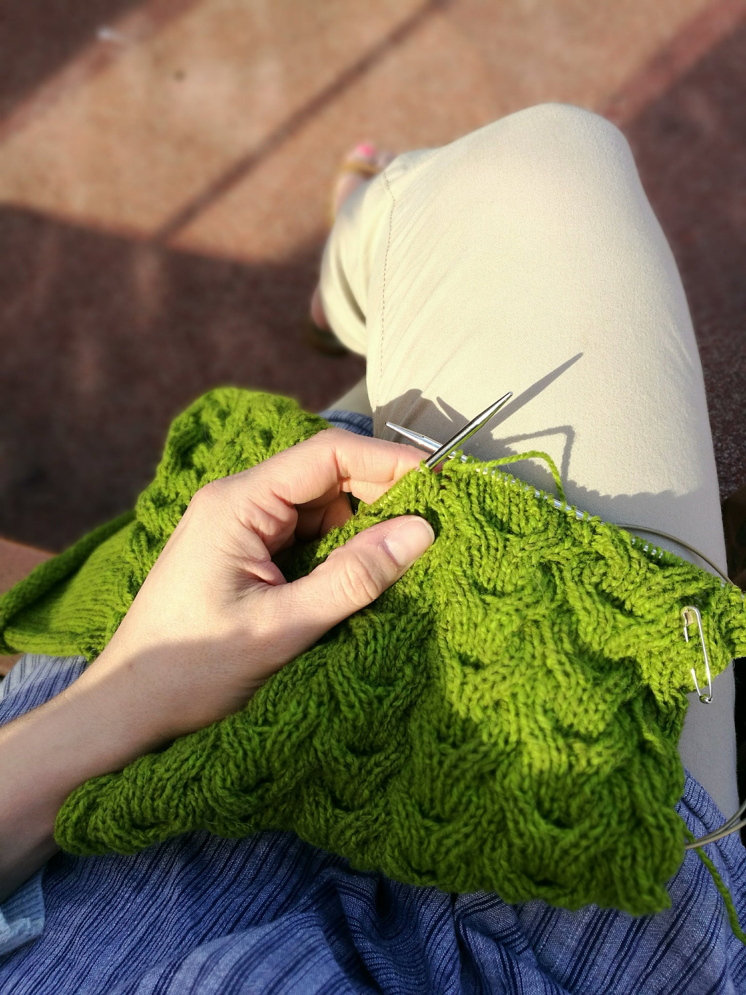 Image of knitting shot from above, in warm sunshine. A white skinned hand holds the needle, the project is a cabled cowl in green yarn.